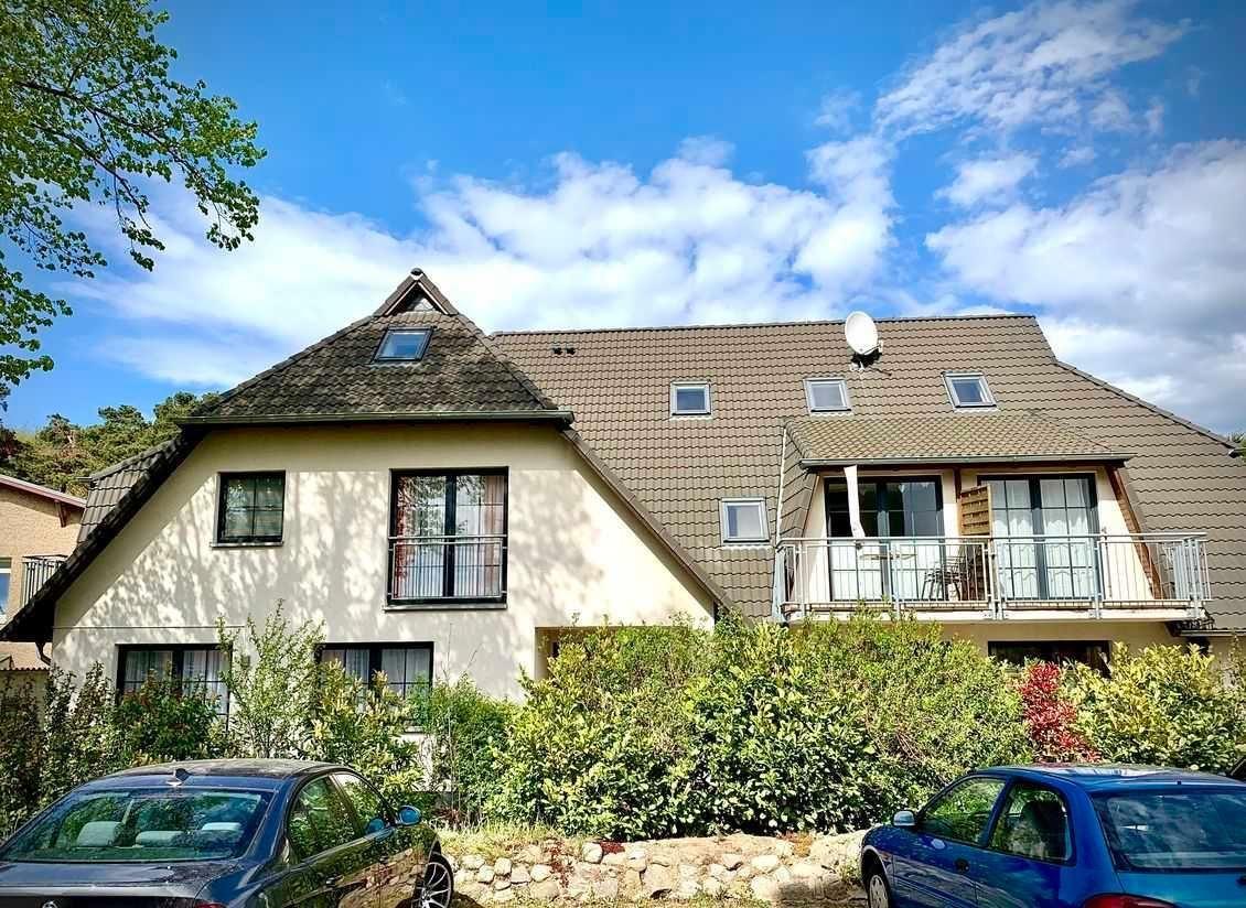 Ein zweistöckiges Haus mit Balkon, Dachfenstern und einem Satellitenschüssel. Vor dem Haus stehen zwei Autos.