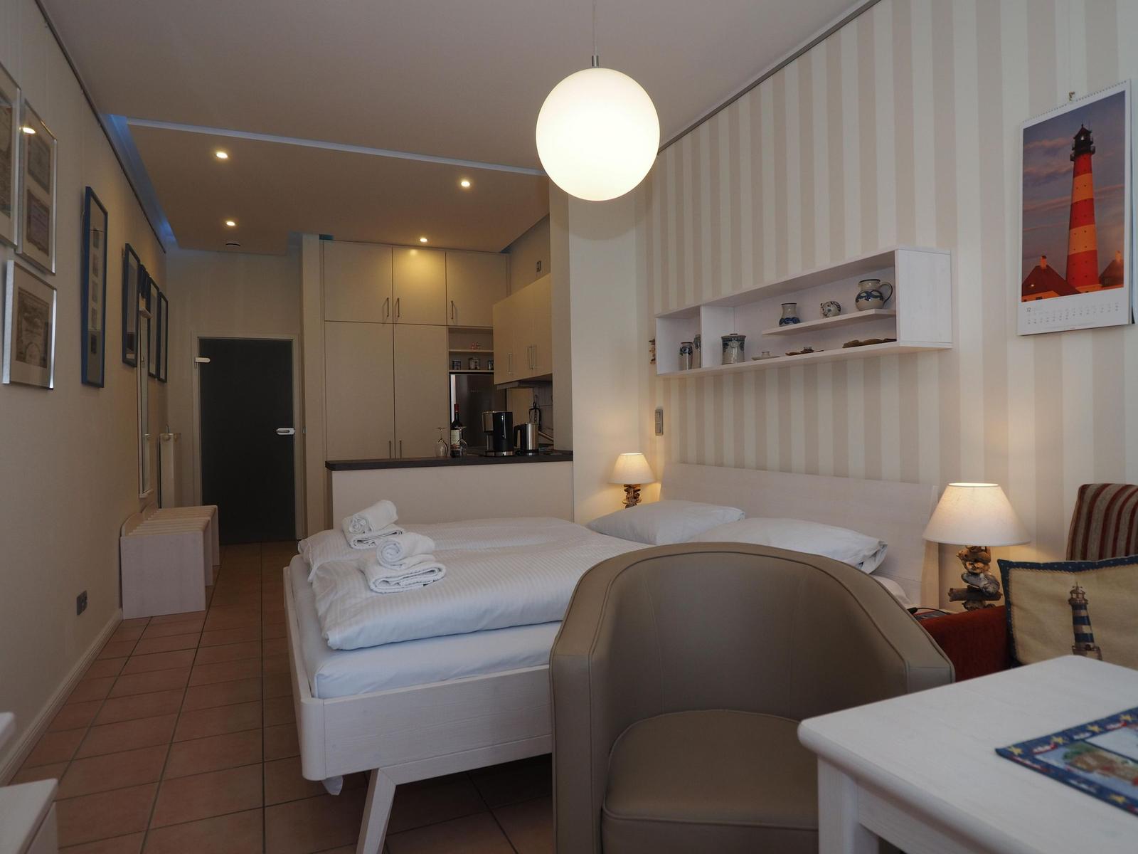 Schlafzimmer mit Bett, Schreibtisch und Küchenbereich im Hintergrund.