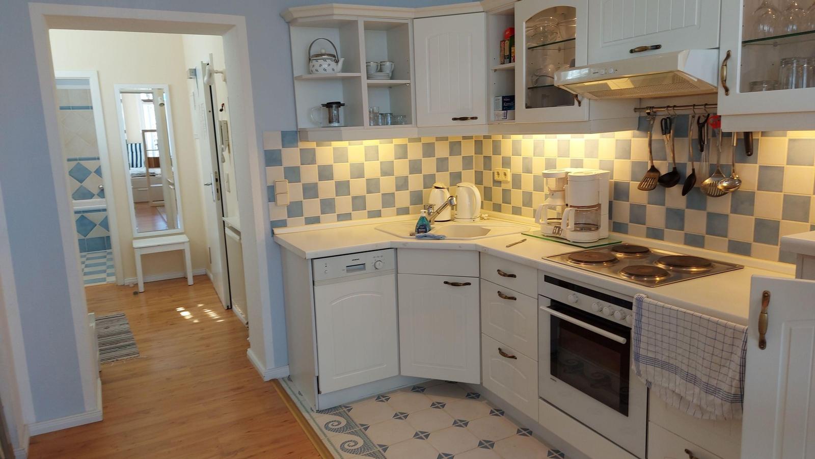 Küche mit weißen Schränken, Spüle, Kochfeld und Backofen. Fliesenwand und Holzboden.
