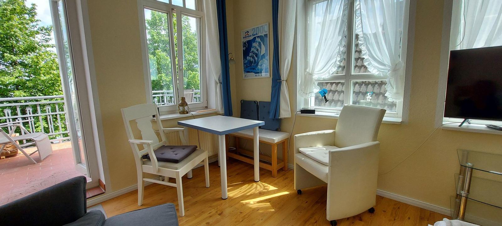 Ein Raum mit Tisch, Stühlen und Fernseher. Durch Fenster und Balkontür ist eine Terrasse sichtbar.