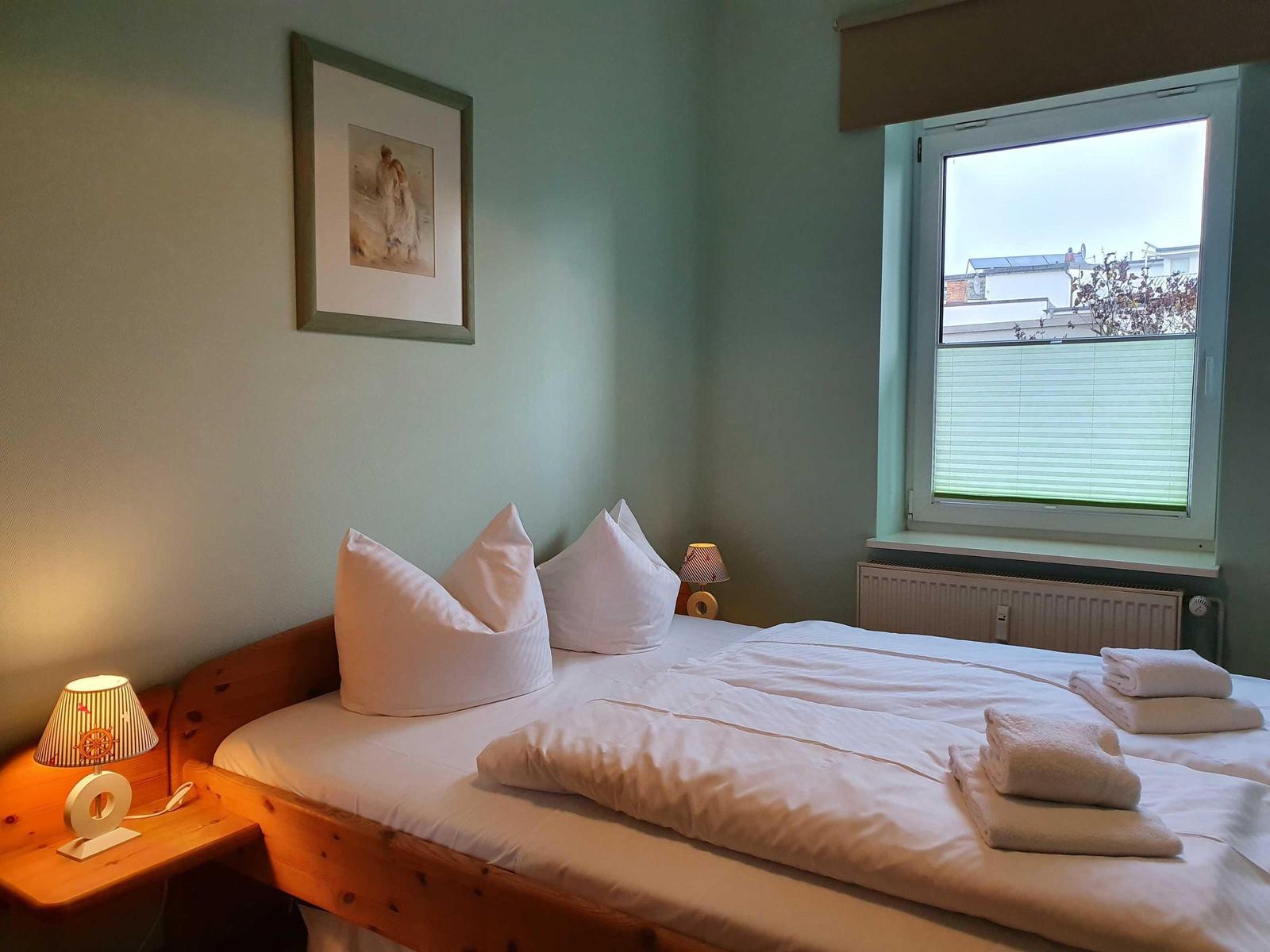 Ein Schlafzimmer mit Bett, Nachttisch, Lampe und Fenster. Auf dem Bett liegen Handtücher.