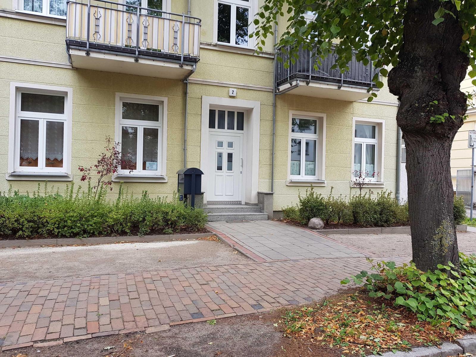 Haus mit Eingang, Balkonen und gepflastertem Weg. Grünflächen und Baum im Vorgarten.