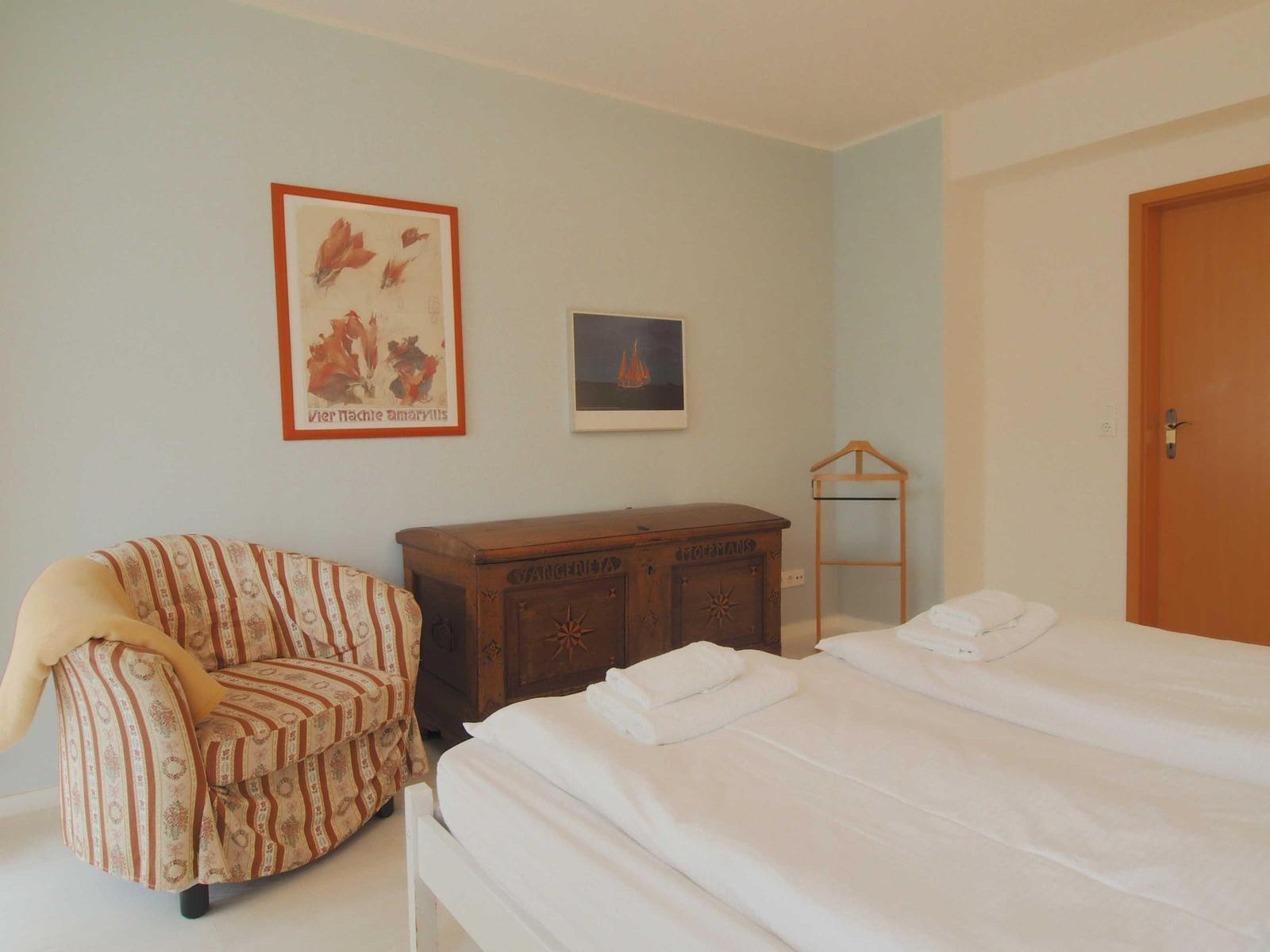 Schlafzimmer mit Bett, Sessel und Holztruhe. An den Wänden hängen zwei Bilder.