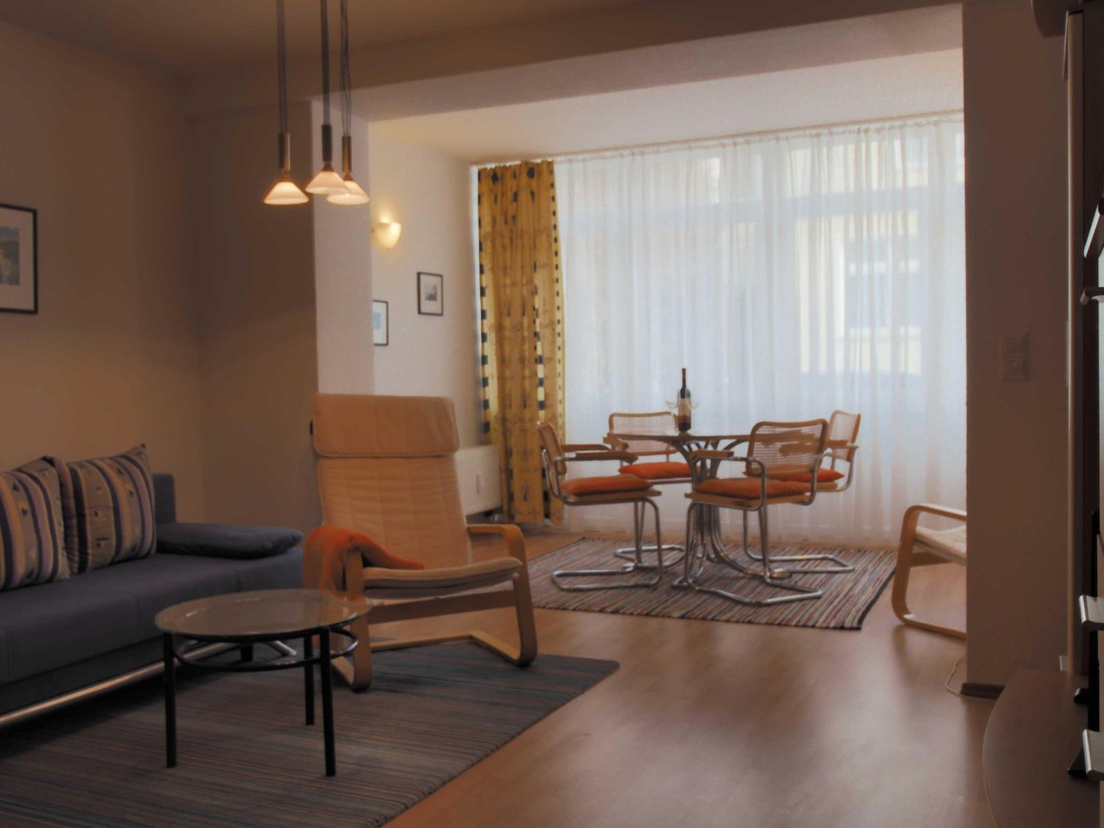 Wohn- und Essbereich mit Sofa, Sessel und Tisch. Helle Fenster mit weißen Vorhängen.