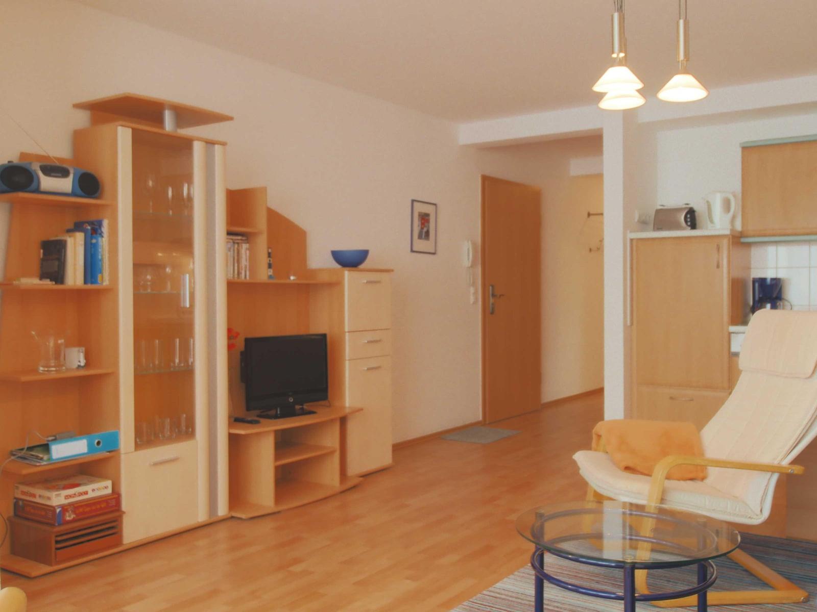 Wohnzimmer mit Holzmöbeln, Fernseher und Sessel. Küche im Hintergrund.