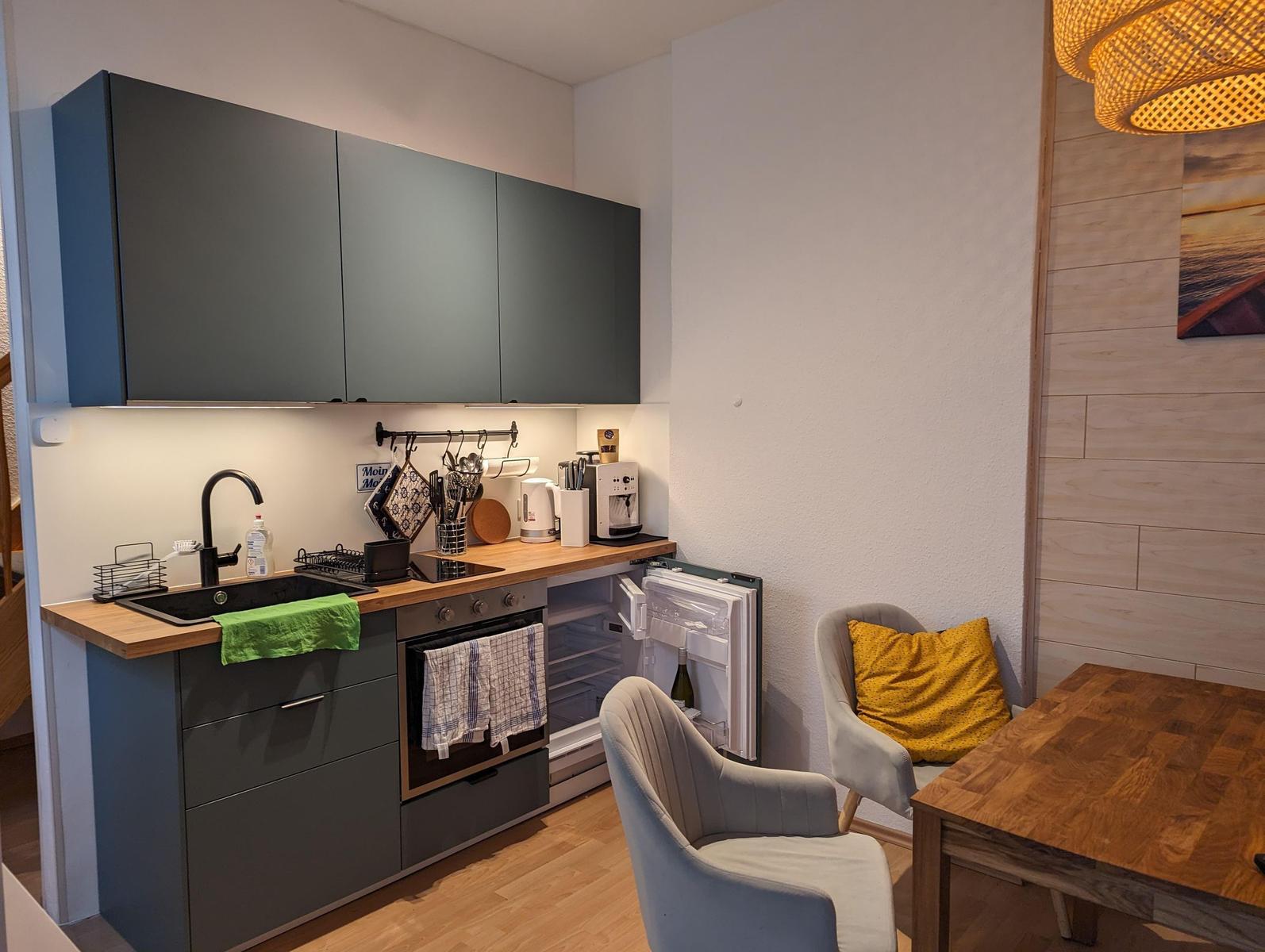 Küche mit Dunstabzug, Spüle und Kühlschrank. Nebenbei Sitzgelegenheit mit Tisch.
