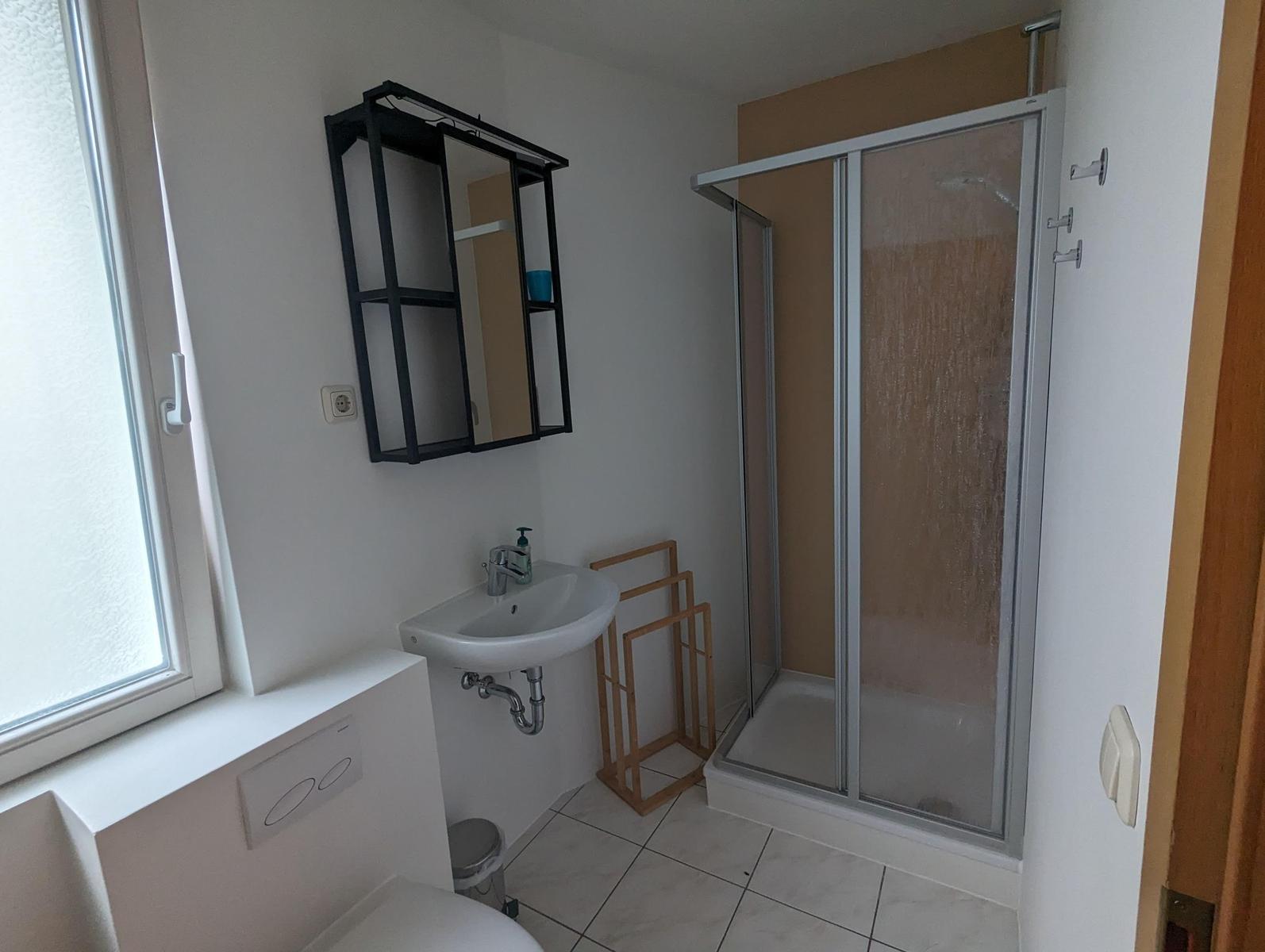 Badezimmer mit Dusche, Waschbecken und Toilette. Fenster sorgt für Tageslicht.
