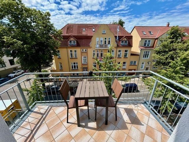 Terrasse mit Tisch und zwei Stühlen, Blick auf Wohnhäuser und Bäume unter blauem Himmel.