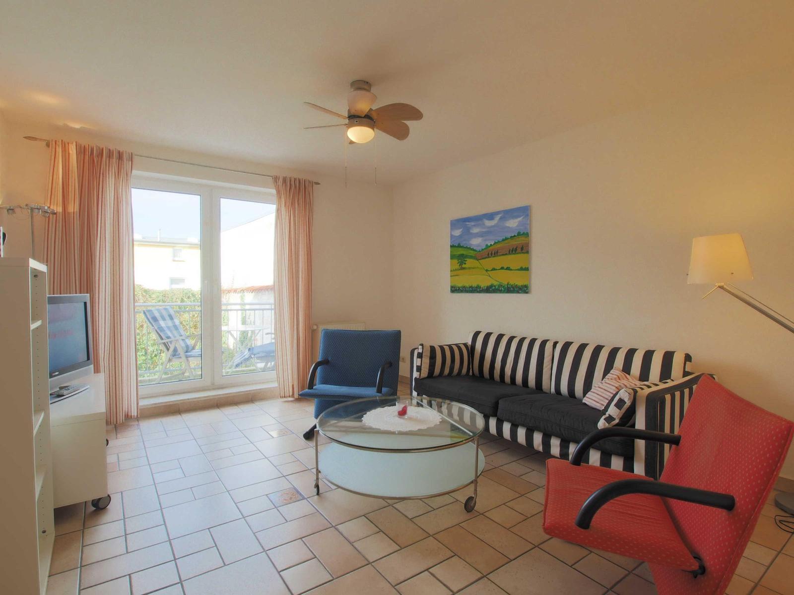Wohnzimmer mit Sofa, Stühlen und TV. Balkon mit Liegestühlen. Wandbild und Deckenventilator.