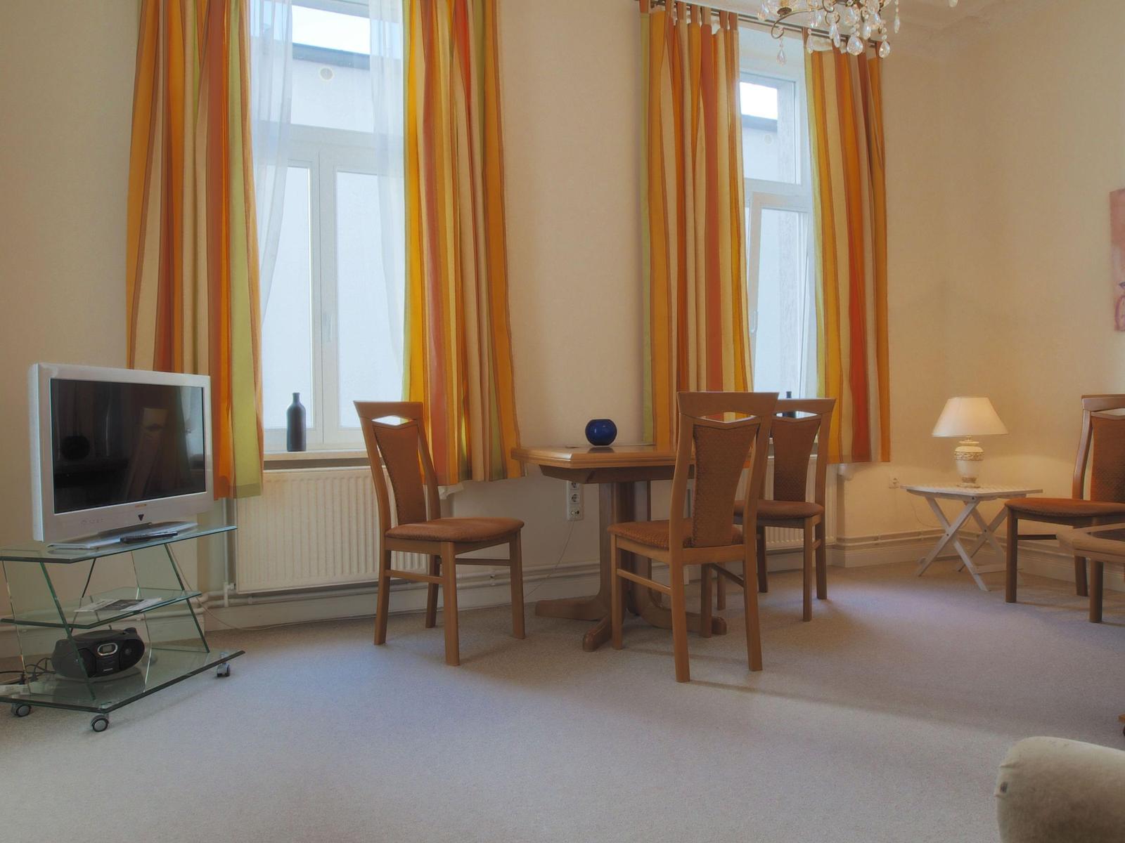 Wohnzimmer mit Esstisch, TV und Fenstern mit bunten Vorhängen.