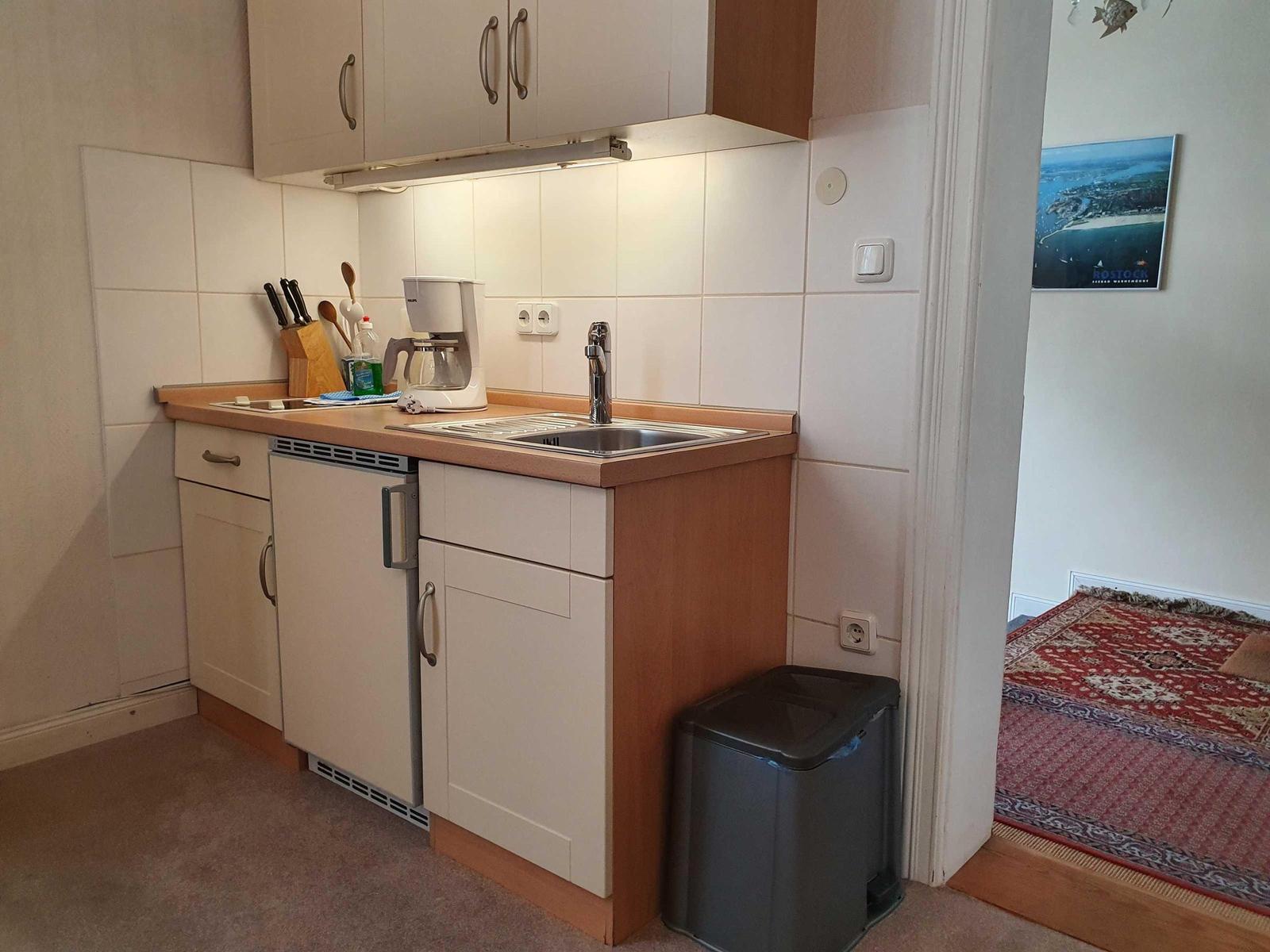 Küche mit Spüle, Kühlschrank und Kaffeemaschine. Blick in Wohnbereich mit Teppich.