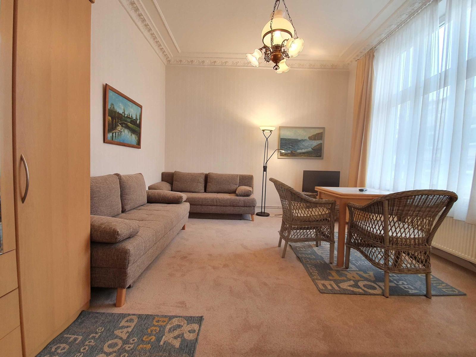 Wohnzimmer mit Sofa, Esstisch und Fenster. Deckenleuchte und Wandbilder.