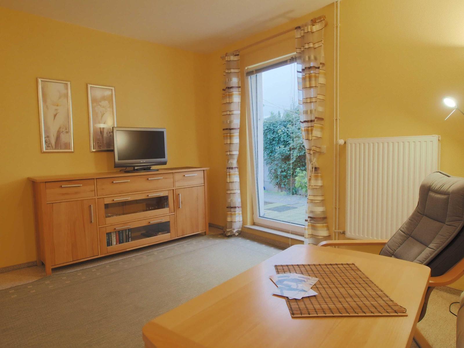 Wohnzimmer mit Fernseher, Tisch und Sessel. Fenster mit Vorhängen. Wand mit Bildern.