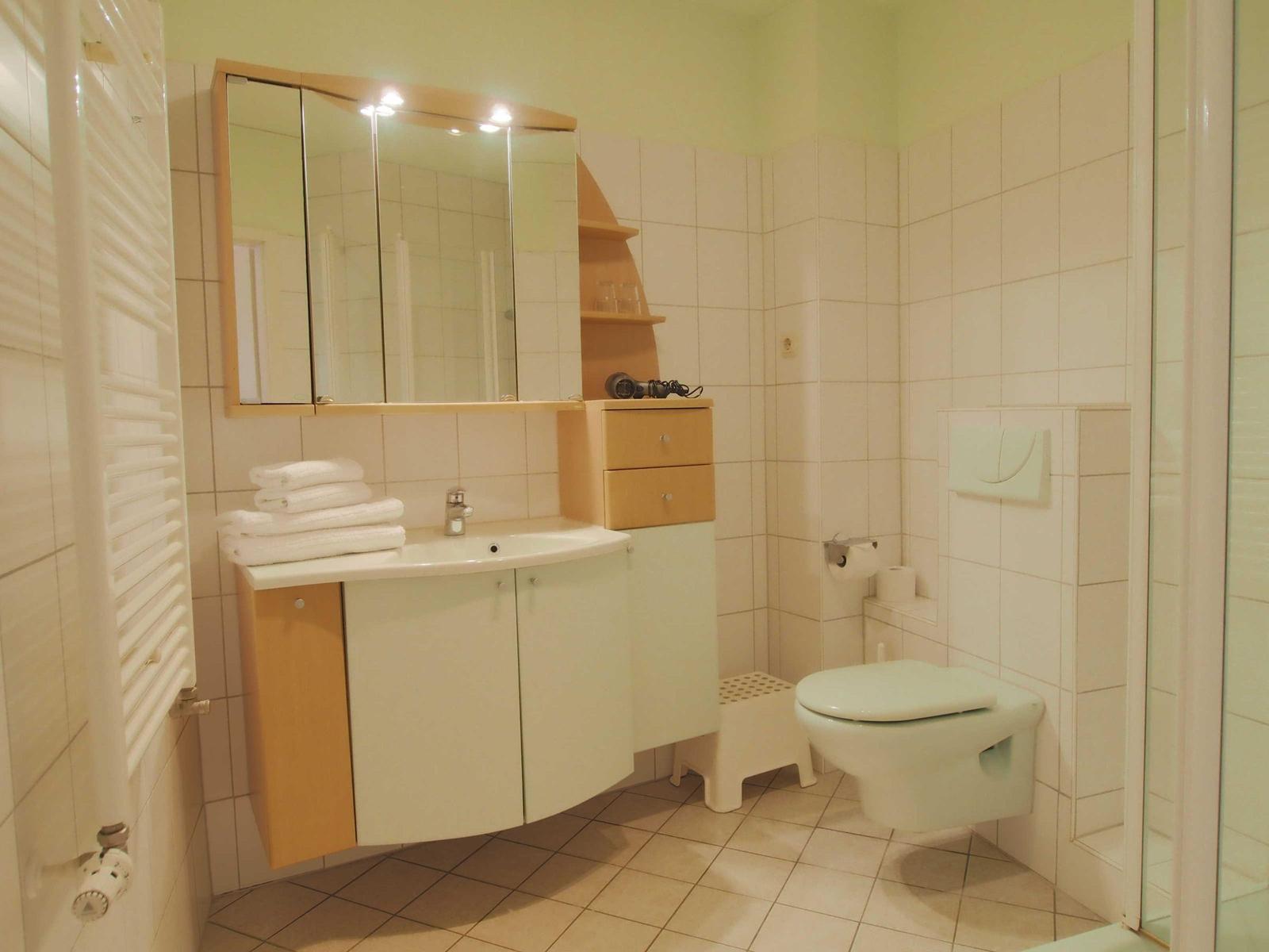Badezimmer mit Waschbecken, Spiegel, Toilette und Dusche. Weiße Fliesen und Holzelemente.
