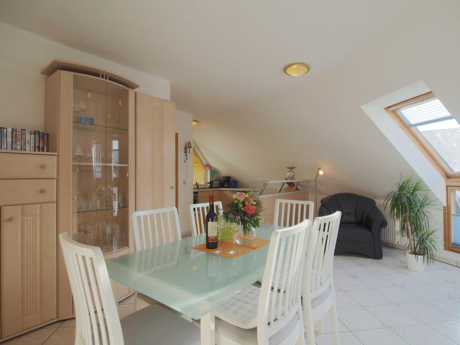 Esszimmer mit Glas-Tisch, weißen Stühlen und Schrank. Dachfenster und Sessel im Hintergrund.