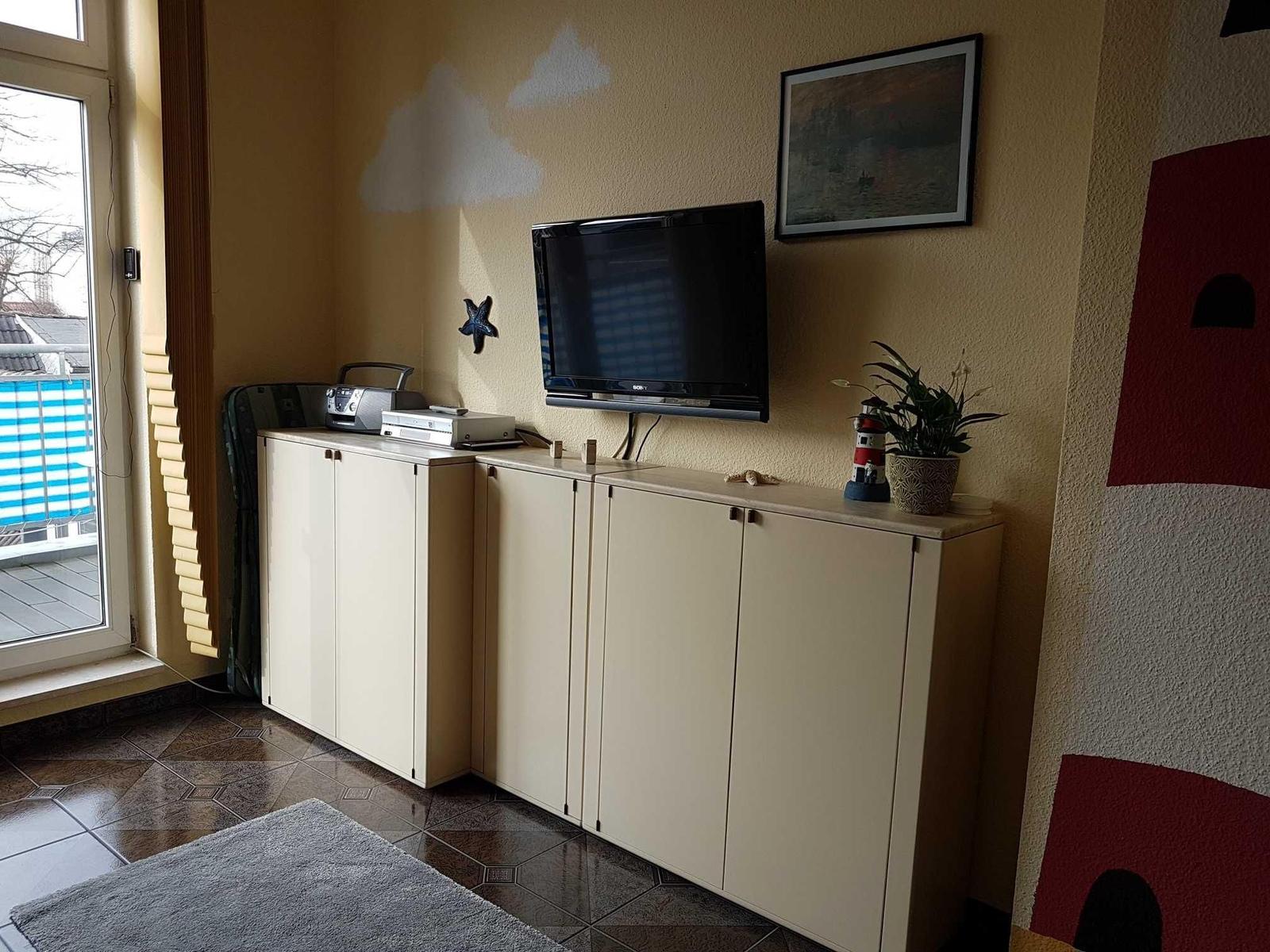TV auf Möbelwand, daneben Pflanze und Leuchtturmfigur. Terrasse mit Blick draußen.