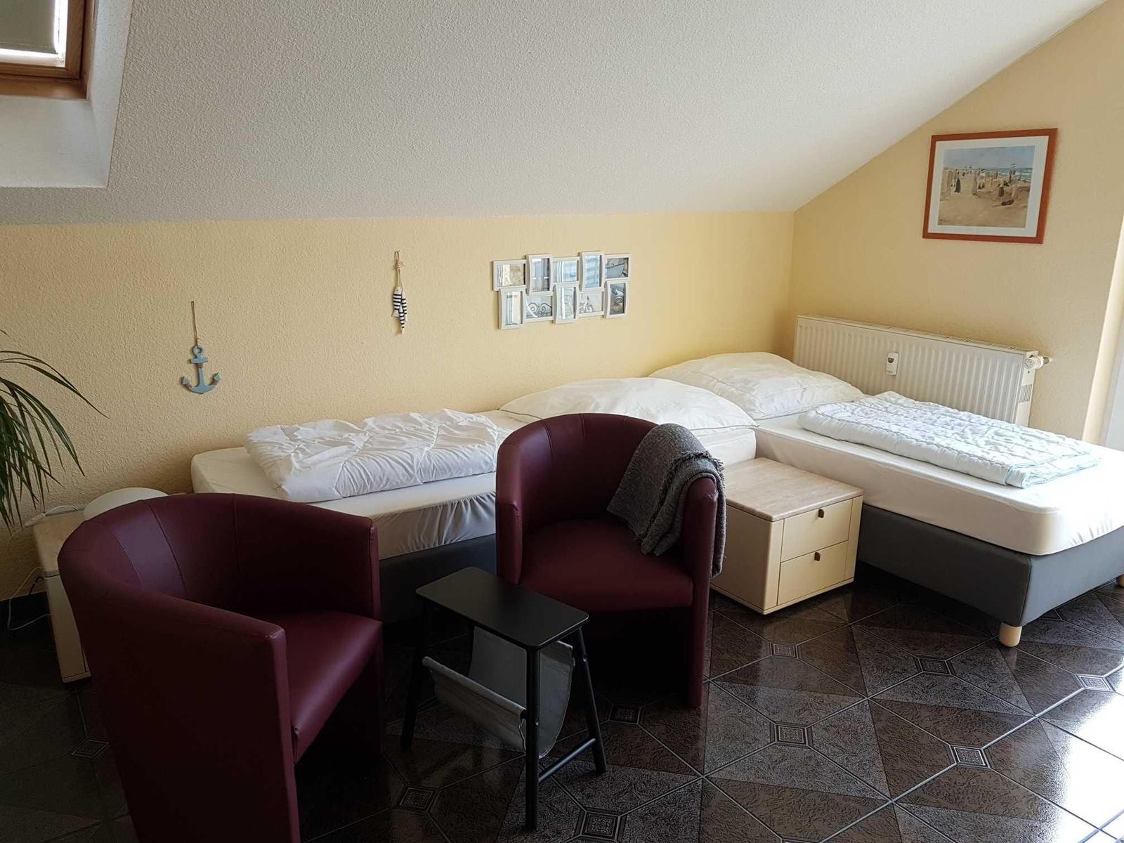 Zimmer mit zwei Betten, zwei roten Sesseln und einem Schrank. Wanddekoration und Fenster sichtbar.