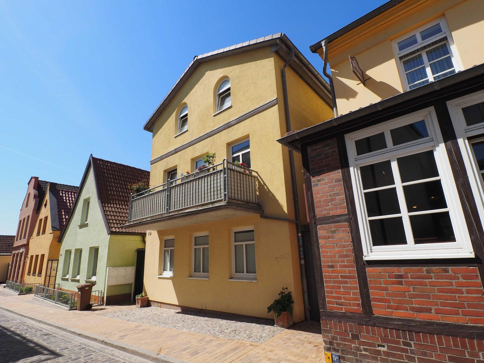 Gelbes Haus mit Balkon und Fenstern an einer Straße mit gepflastertem Gehweg.