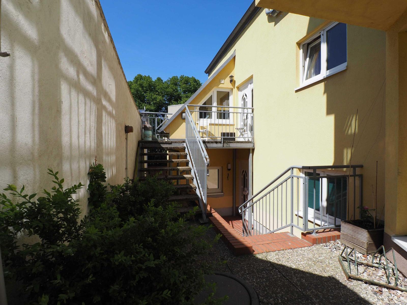 Hinterhof mit Treppe und Balkon an gelbem Haus mit Pflanzen und Steinplatten.
