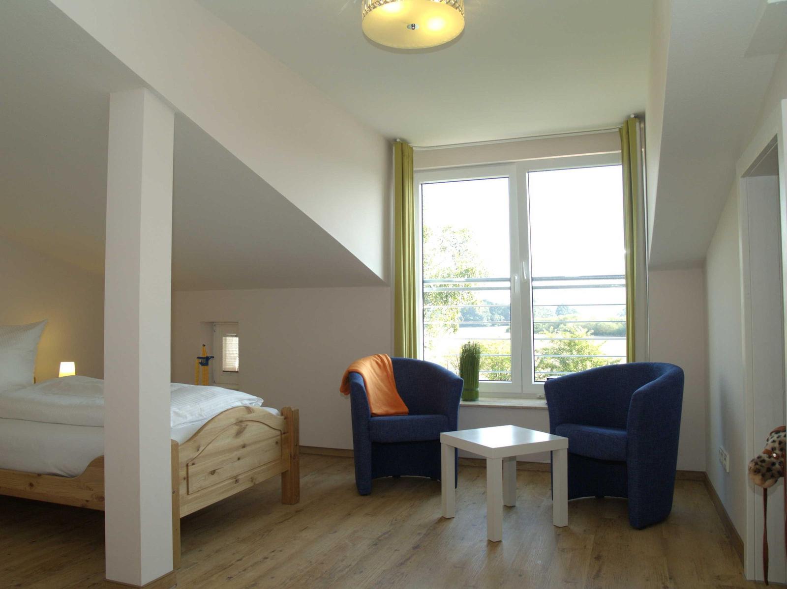 Schlafzimmer mit Doppelbett, zwei blauen Sesseln und großem Fenster mit Blick.