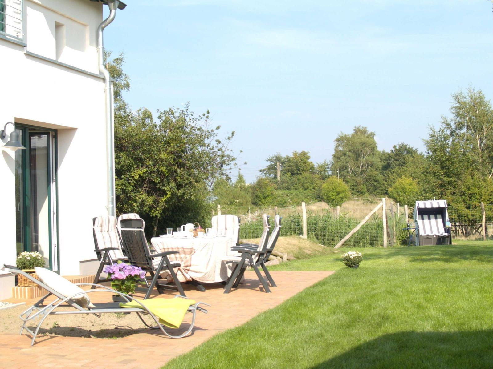 Terrasse mit Tisch, Stühlen und Liege. Grasfläche und Strandkorb im Hintergrund.