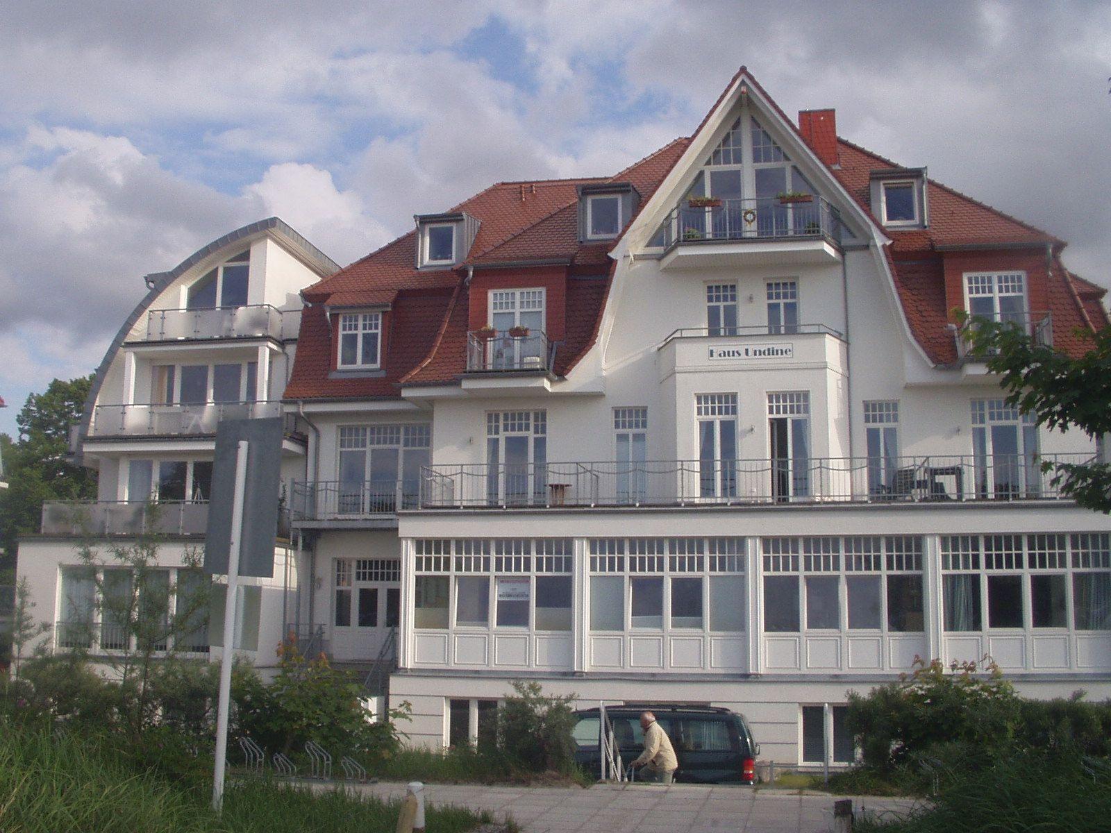Wohnhaus mit rotem Dach und weißen Fassaden, mehrere Balkone und Fensterfronten.