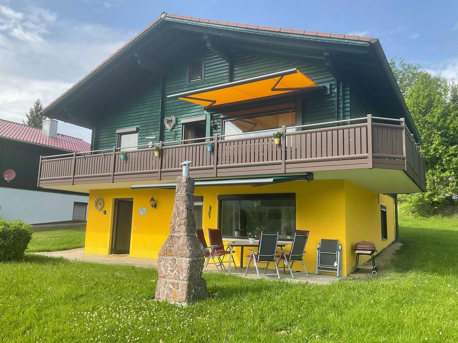 Two-story house with balcony, grill, and garden. Yellow lower level, green upper level.