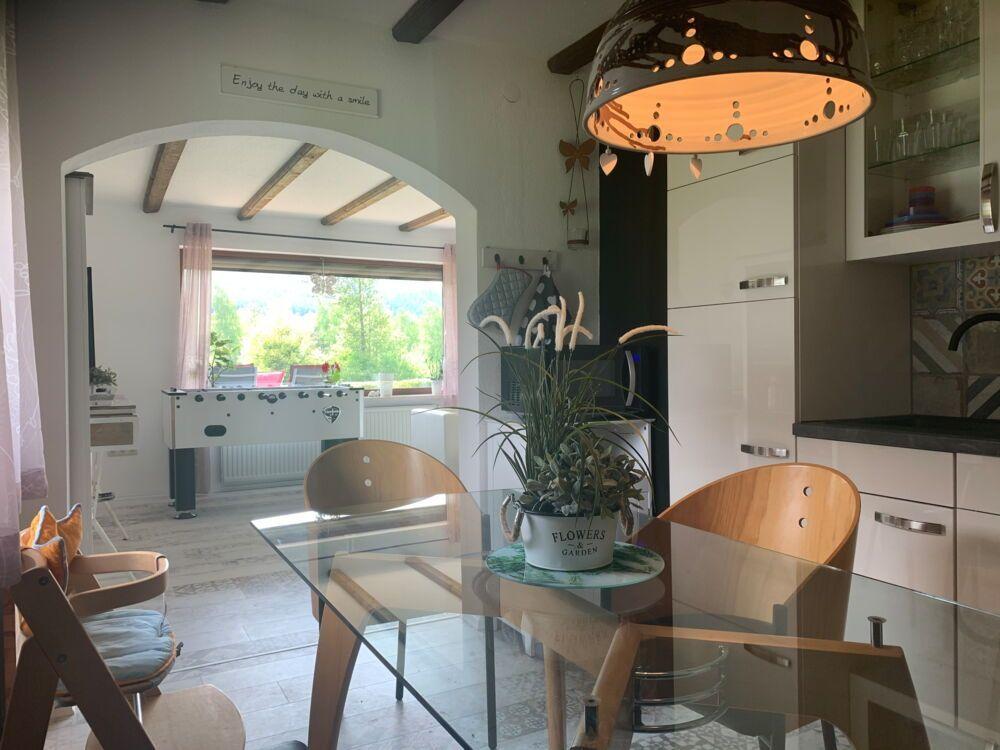 Dining area with glass table and wooden chairs. Kitchen and living area with view of green landscape.