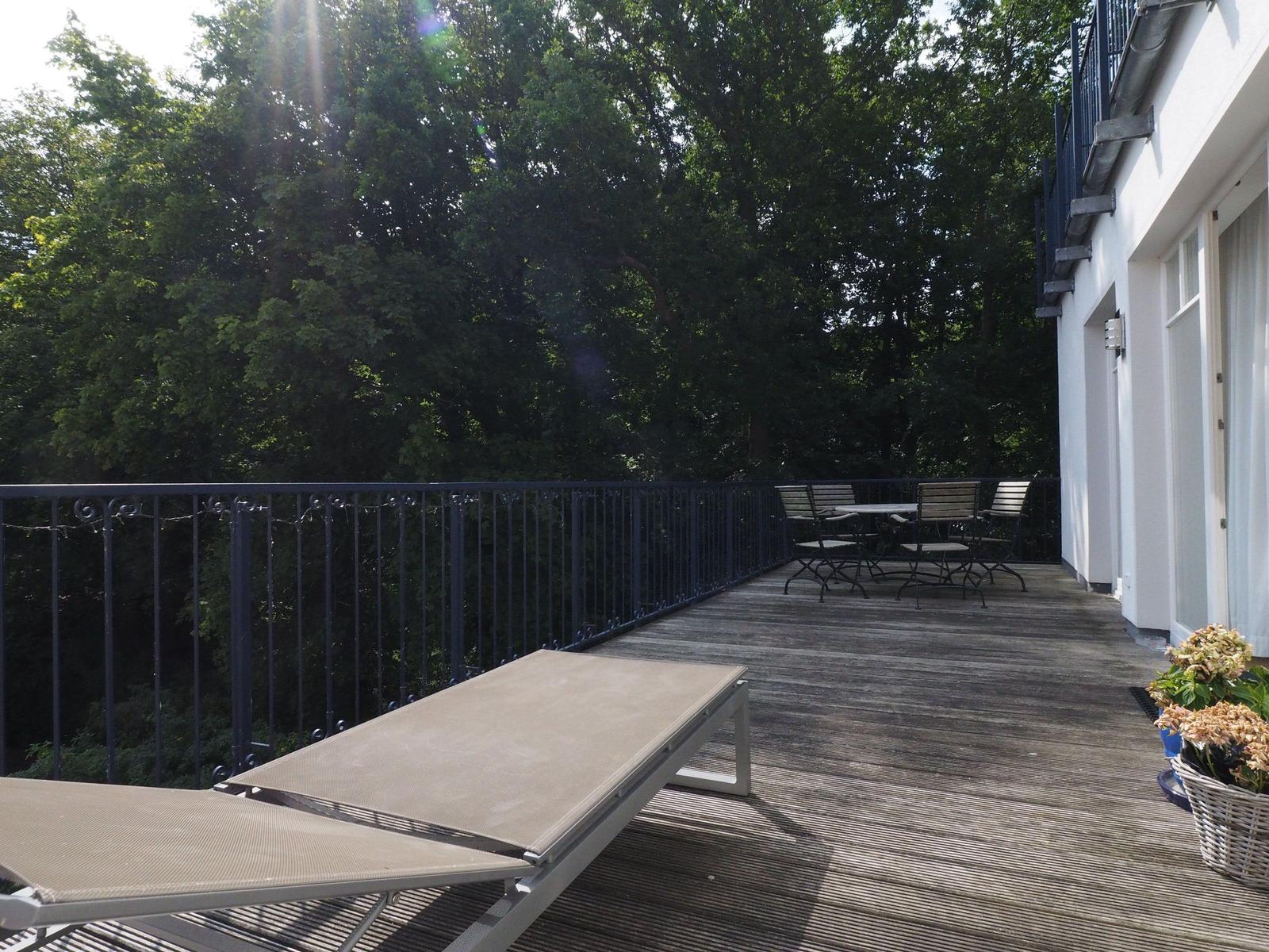 Terrasse mit Liegestuhl und Tischset vor grüner Baumreihe