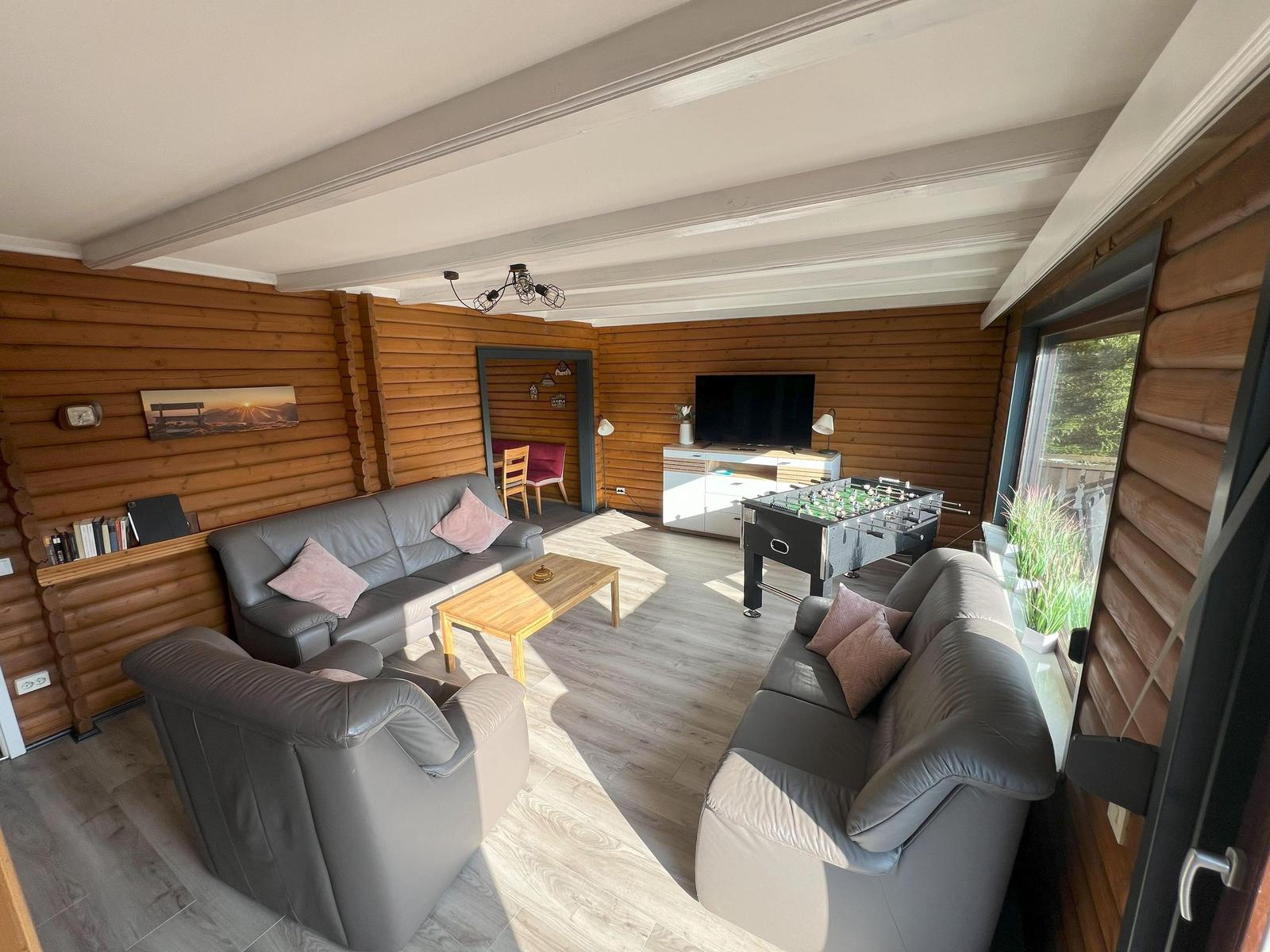 Living room with sofa, armchair, table, and foosball table. Wooden walls and window view.