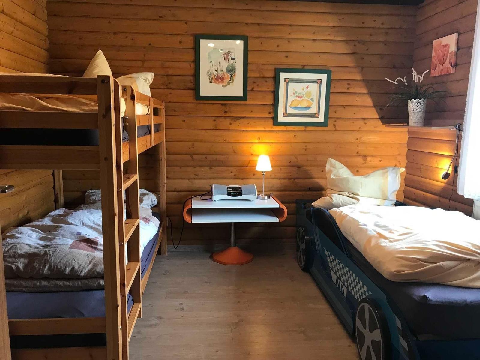 Room with bunk bed and car bed, wooden walls, table with lamp and pictures.