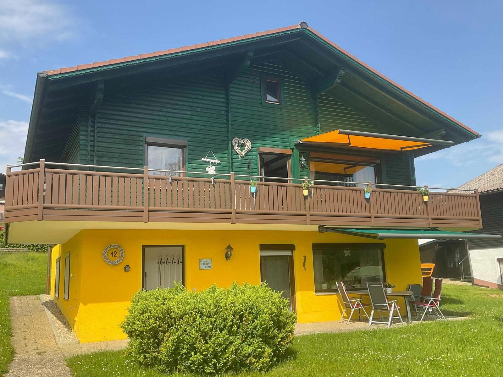 Two-story house with balcony and garden. Ground floor yellow, upper floor green.
