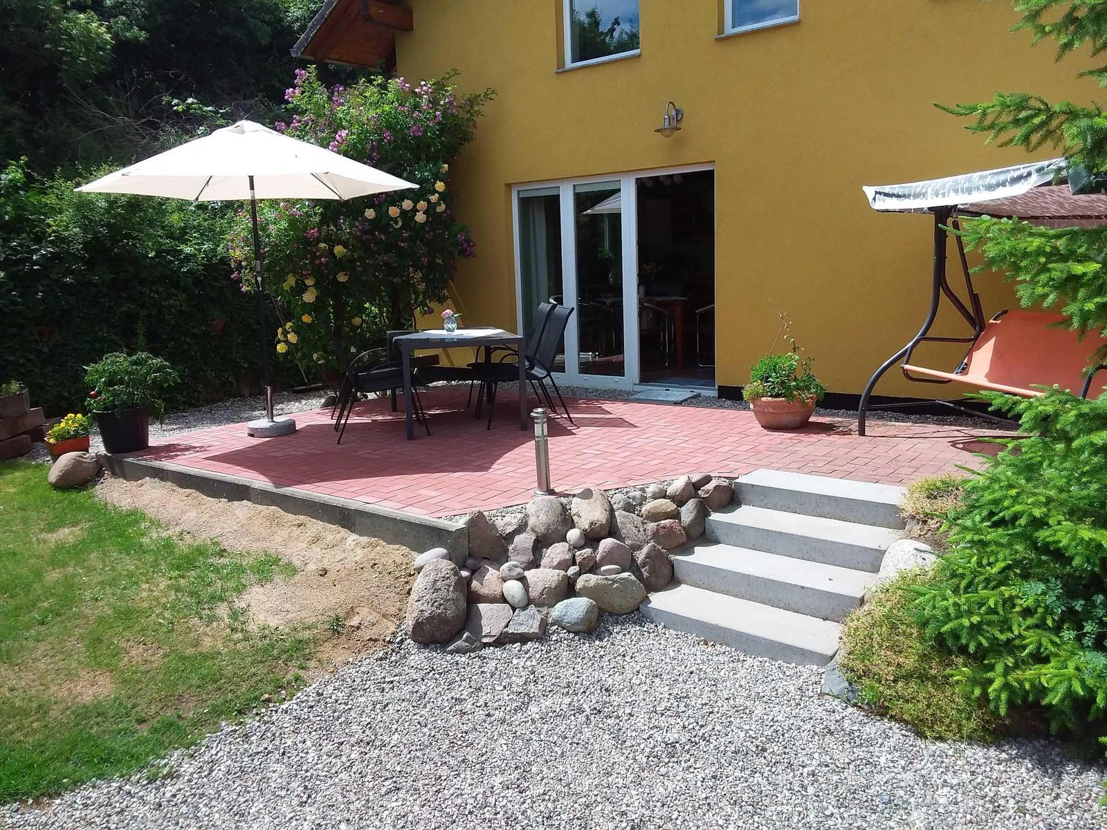 Terrasse mit Tisch, Stühlen und Sonnenschirm. Garten mit Steinstufen und Grasfläche.