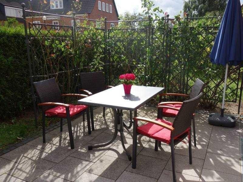 Hinterhof mit Tisch, vier Stühlen und blauem Sonnenschirm. Grünflächen und Zaun im Hintergrund.