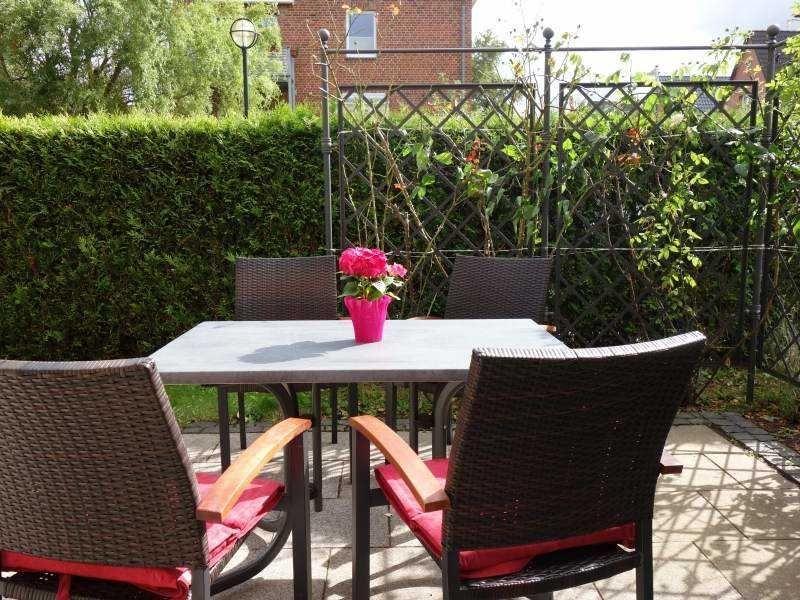 Terrasse mit Tisch und Stühlen, eine Blumenvase und grüne Hecke im Hintergrund.