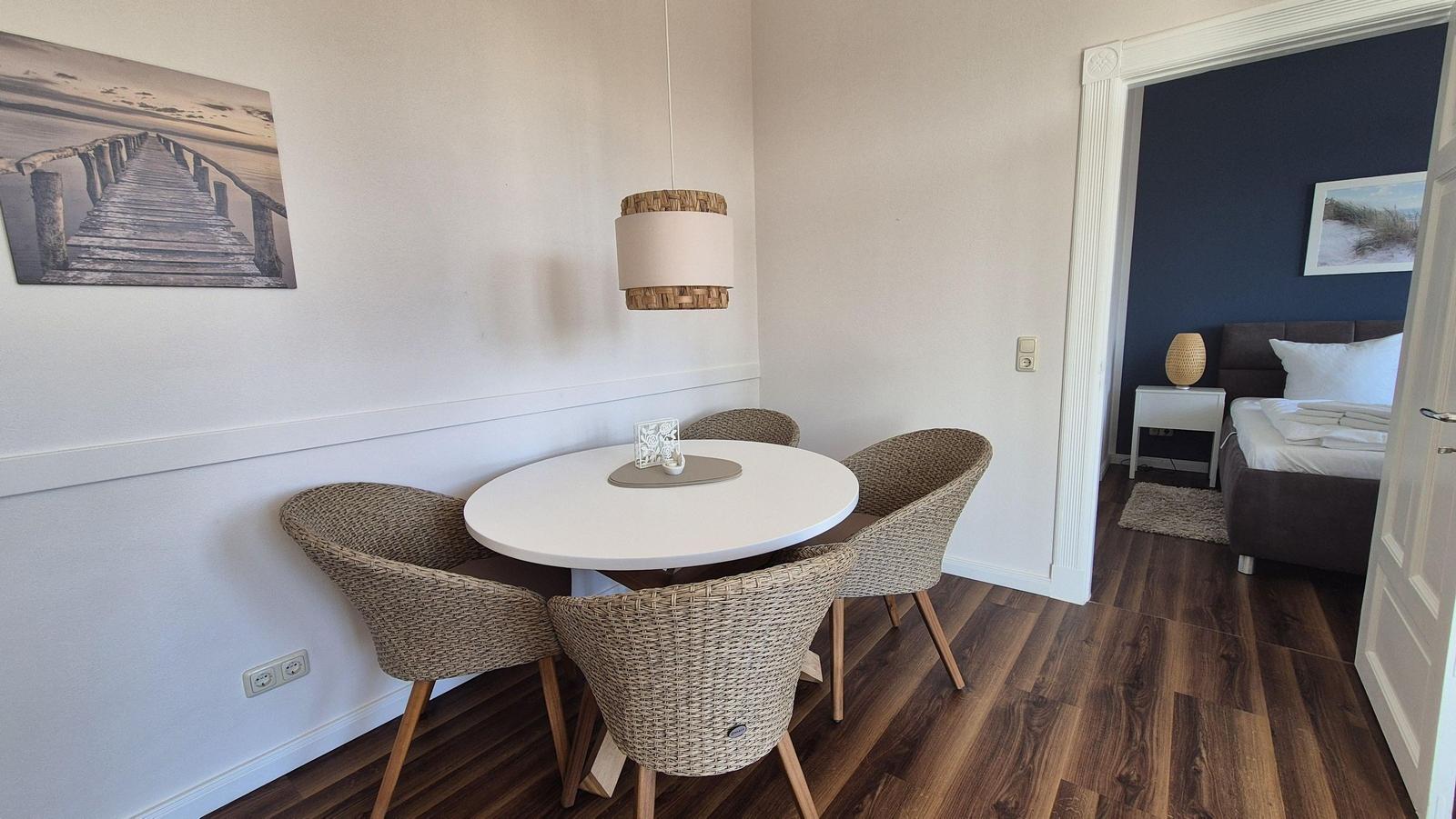 Essbereich mit runden Tisch und vier Stühlen. Im Hintergrund ist das Schlafzimmer mit Bett und Wandbild sichtbar.
