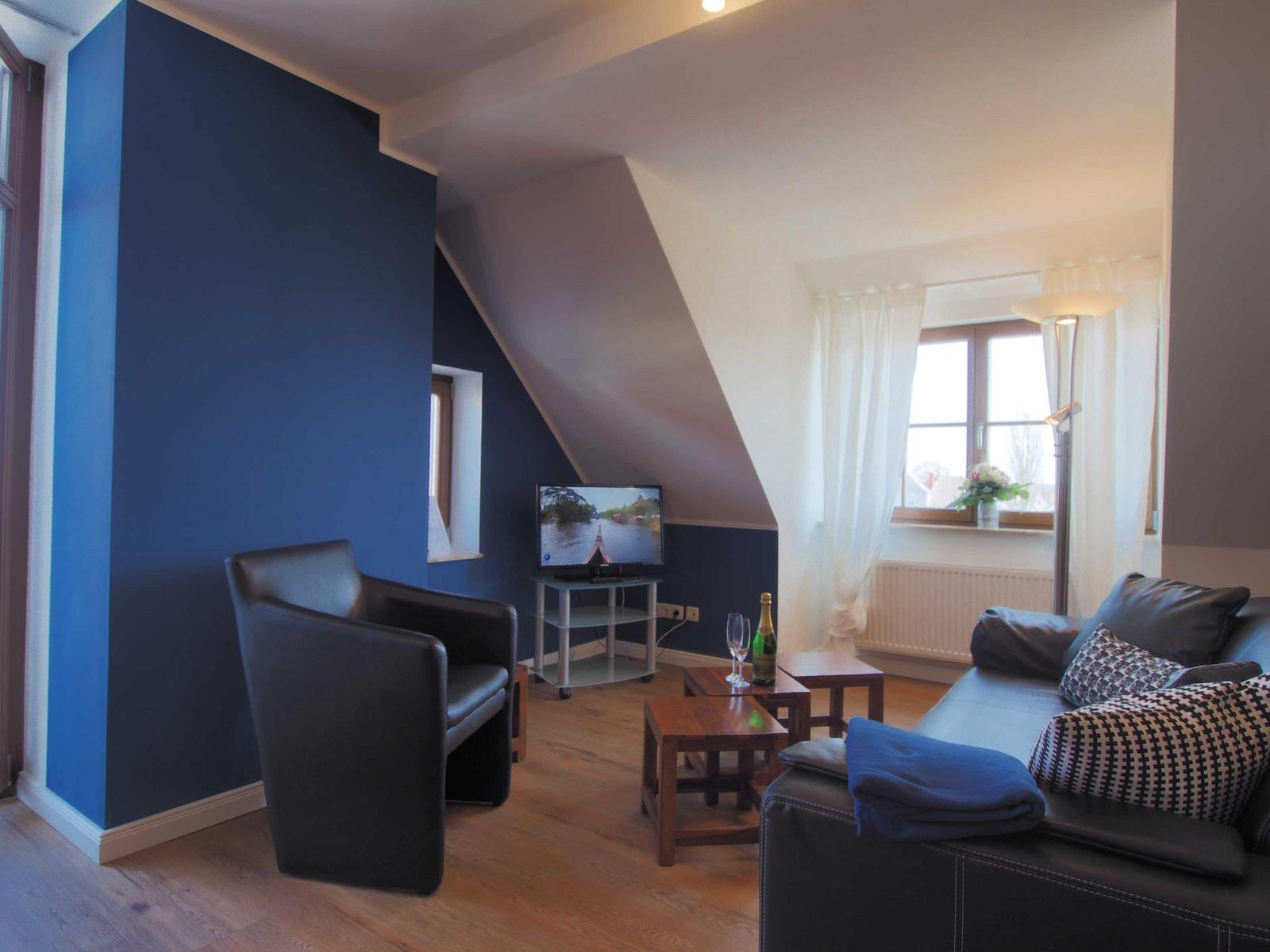 Wohnzimmer mit Sofa, Sessel, Fernseher und Fenster. Wand in Blau und Holzboden.