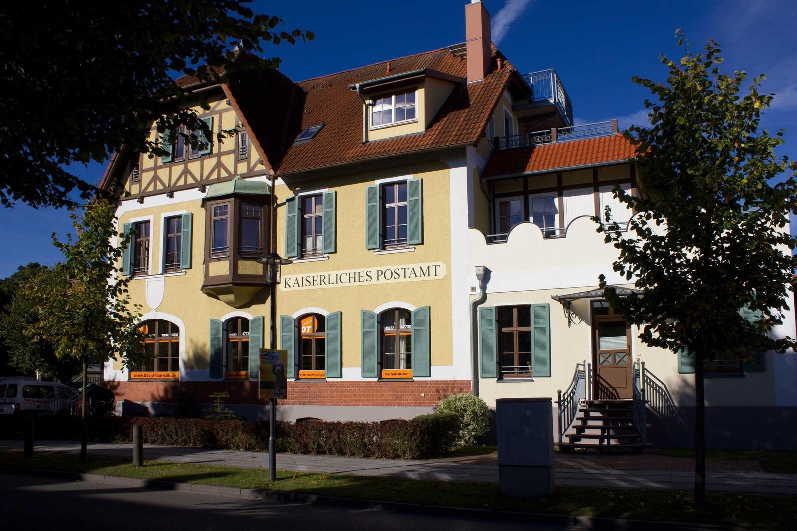 Historisches Gebäude mit Balkon und rotem Dach. Eingang mit Treppe und grünen Fensterläden.