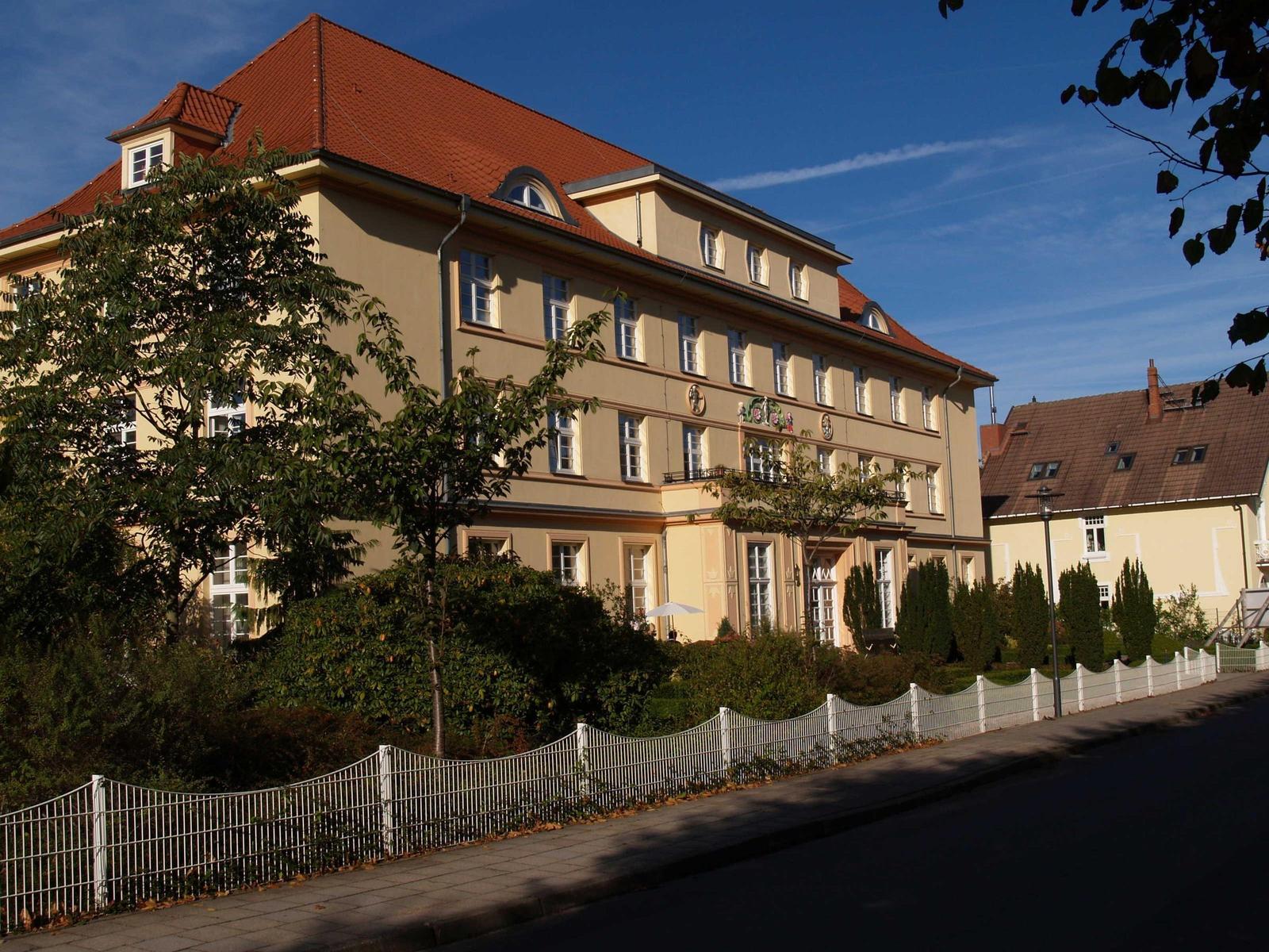 Ein mehrstöckiges Gebäude mit rotem Dach und weißen Fenstern. Vor dem Haus steht ein weißer Zaun.