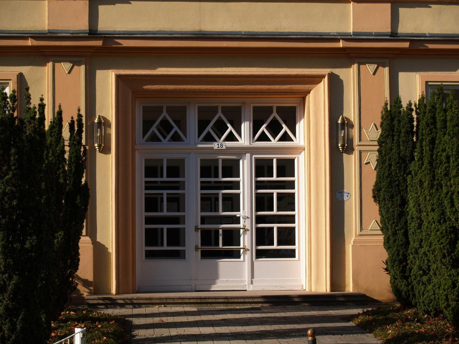 Eingangstür mit weißen Türen und Glasfenstern, Hausnummer 18, flankiert von Nadelbäumen.