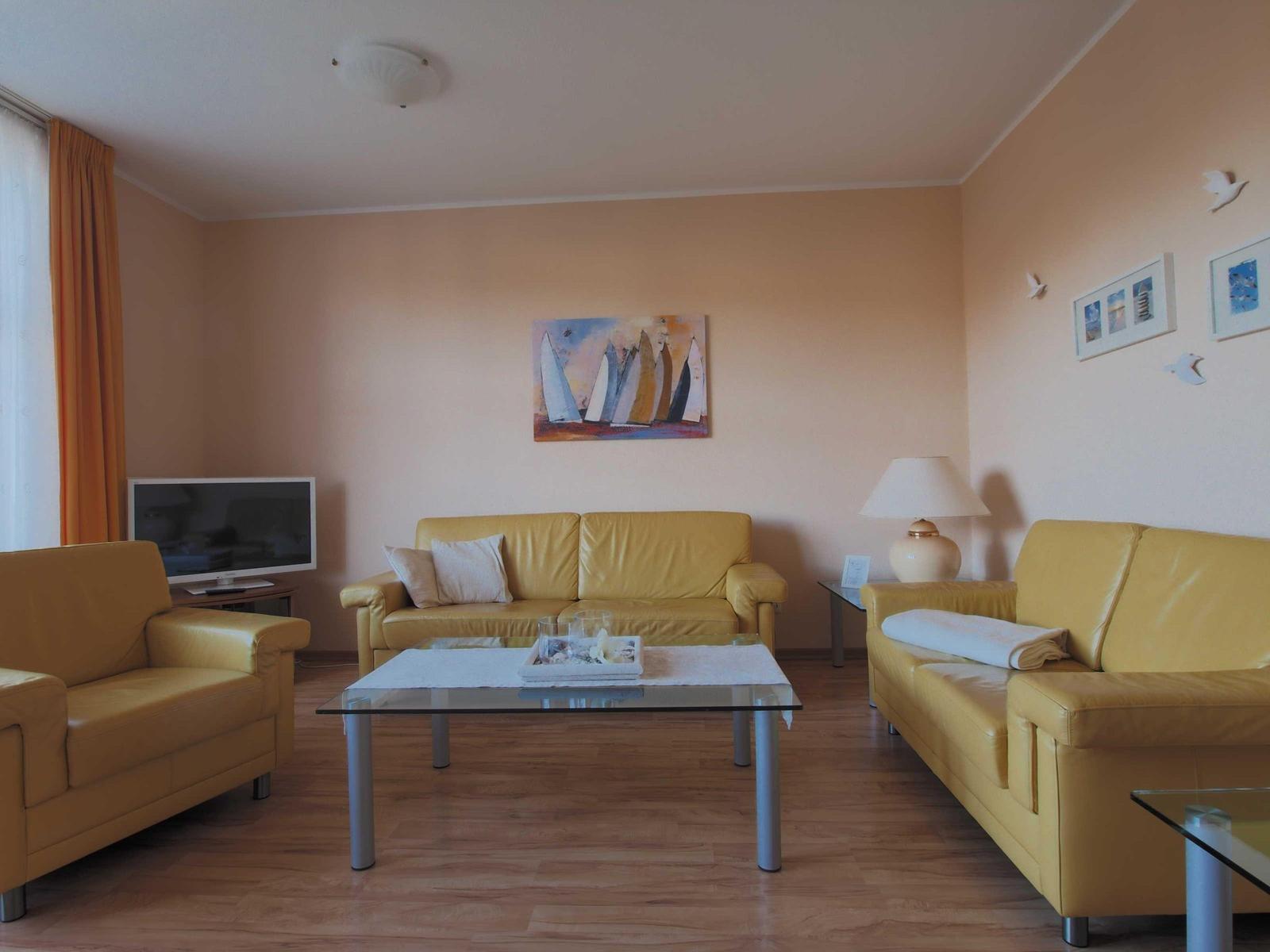 Wohnzimmer mit zwei gelben Sofas, Glas-Tisch und Wandbild mit Segelbooten.