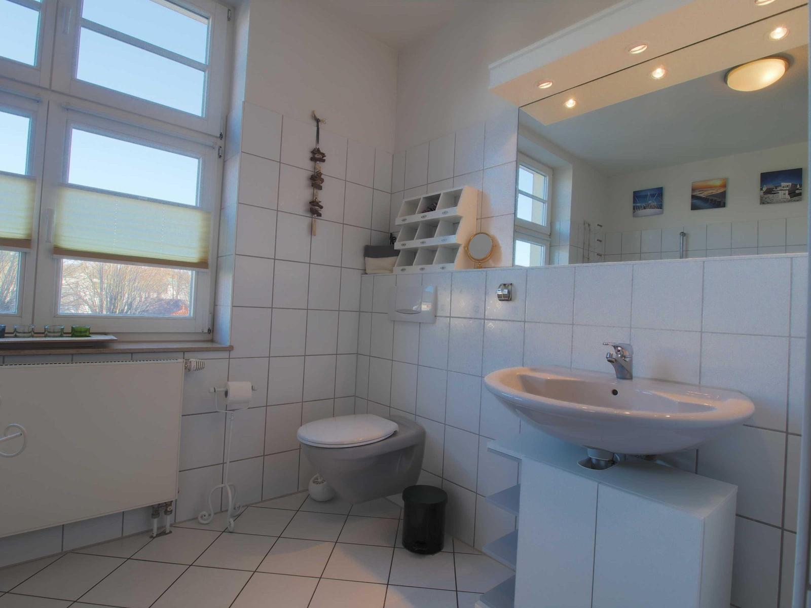 Badezimmer mit Toilette, Waschbecken und Fenster. Weiße Fliesen und Spiegel.