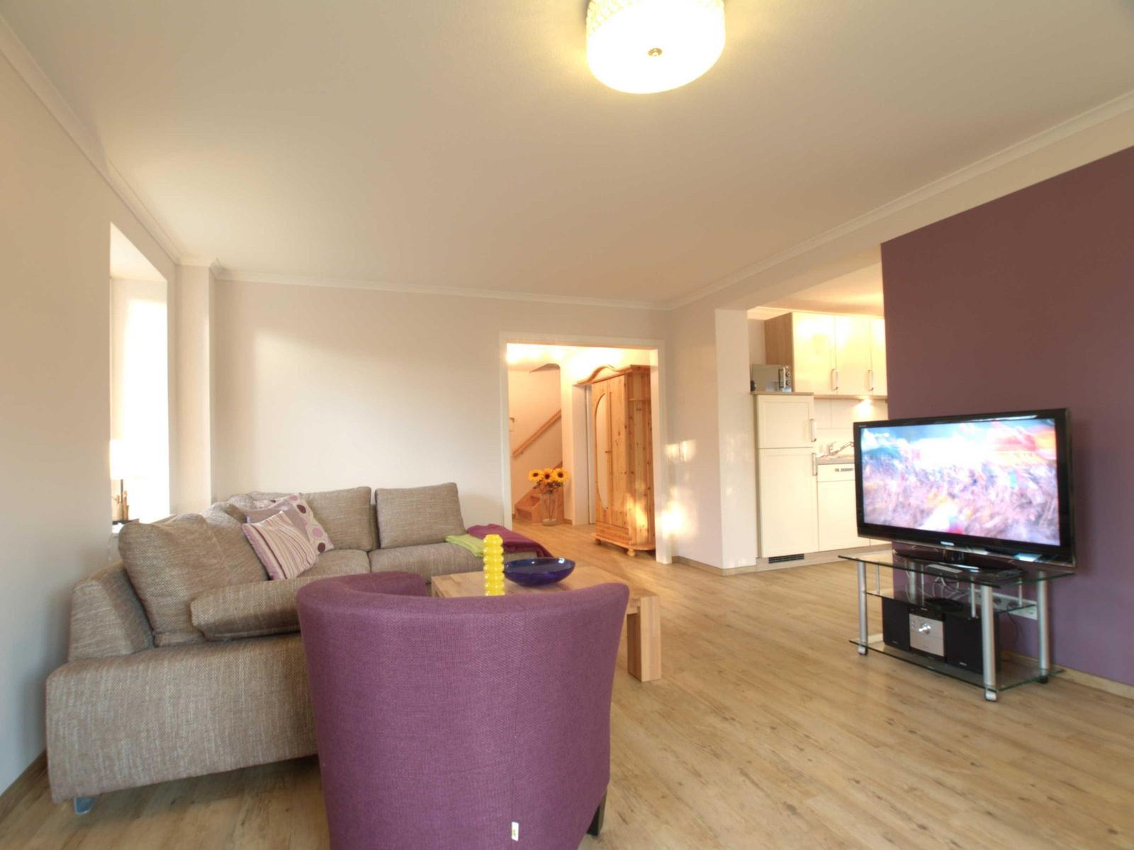 Wohnbereich mit Sofa, Sessel, Fernseher und Küchenbereich im Hintergrund.