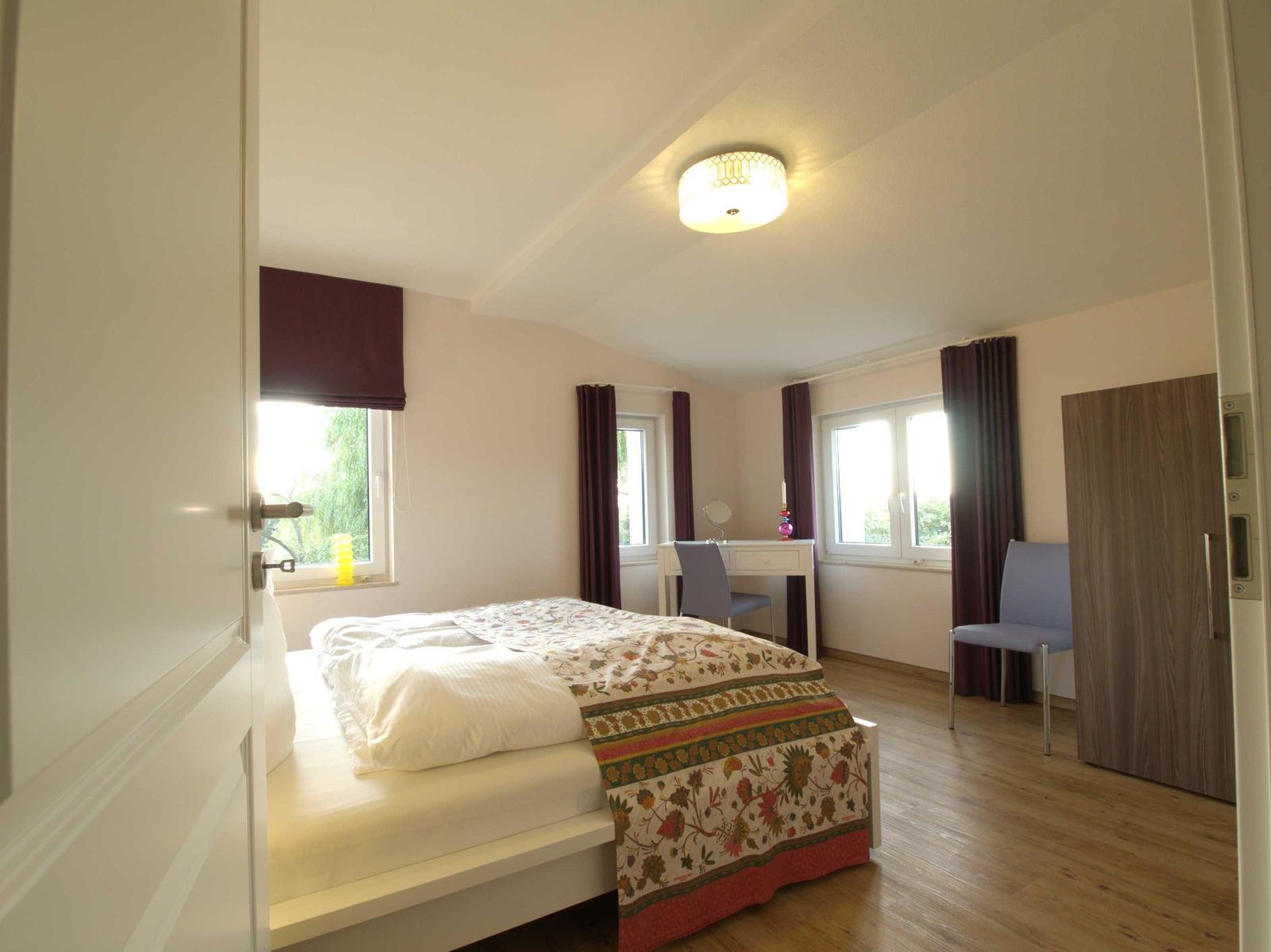 Schlafzimmer mit Bett, Schreibtisch, Stühlen und Fenstern mit Vorhängen.