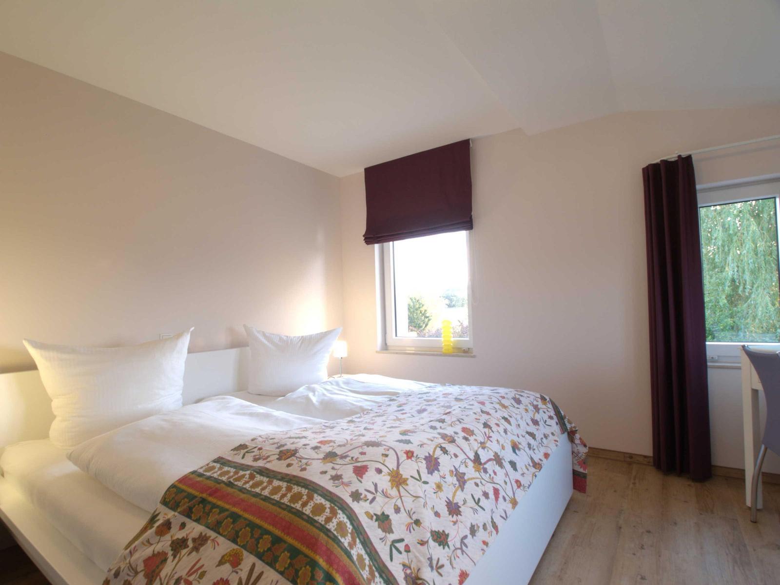 Schlafzimmer mit Bett, Fenster und Vorhängen. Holzboden und weißer Wandschrank.