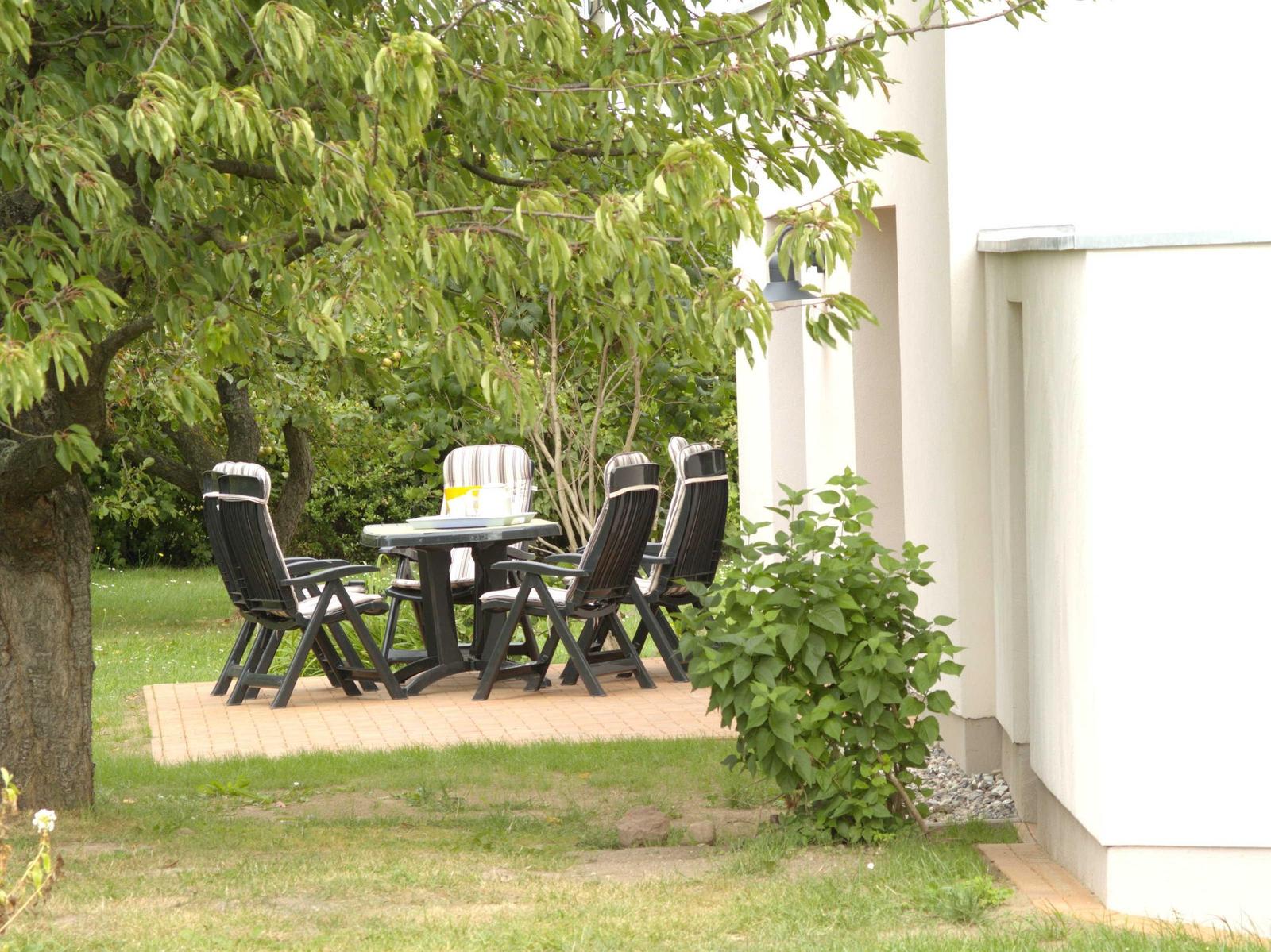 Terrasse mit Tisch und Stühlen unter Bäumen neben weißem Haus