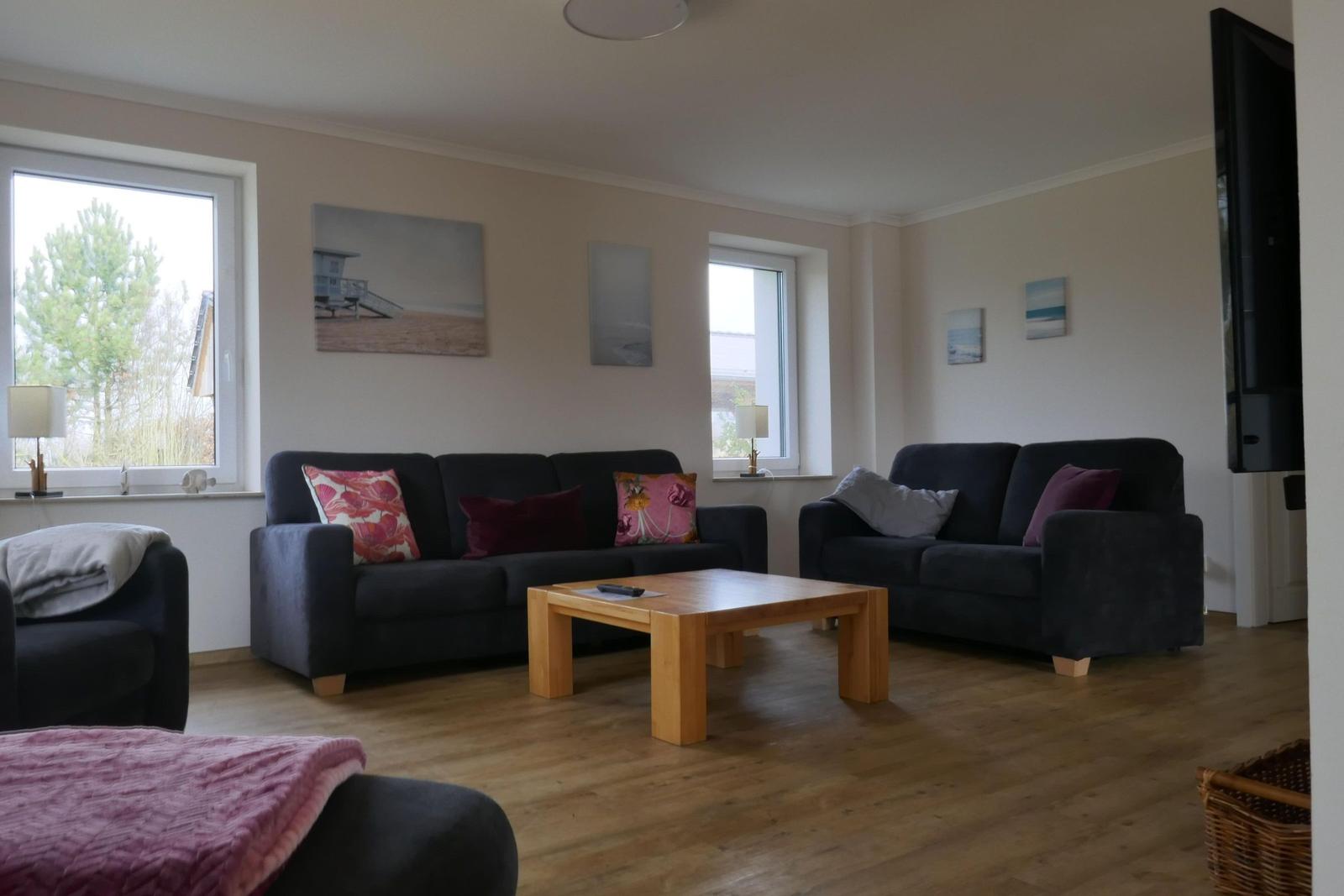 Wohnzimmer mit zwei Sofas, Holztisch und Fenstern. Wandbilder zeigen Strandmotiv.