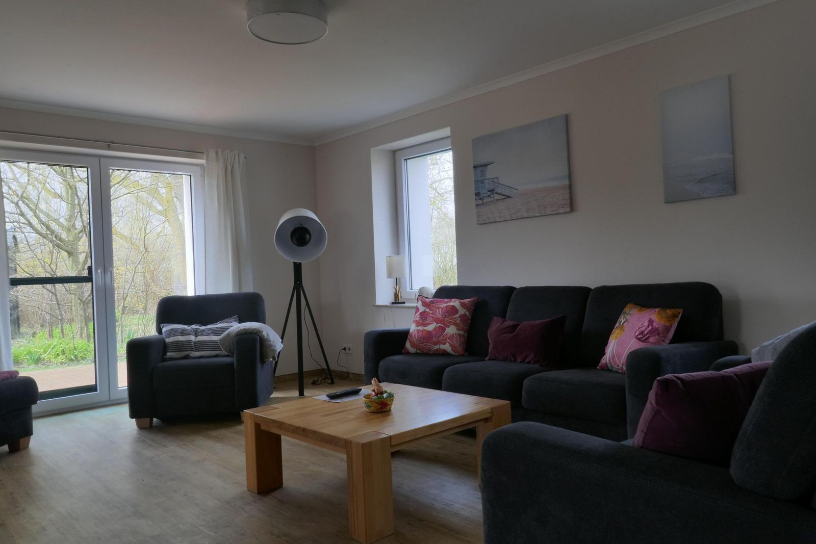 Wohnzimmer mit Sofa, Sessel und Holztisch. Fenster und Wandbilder sichtbar.