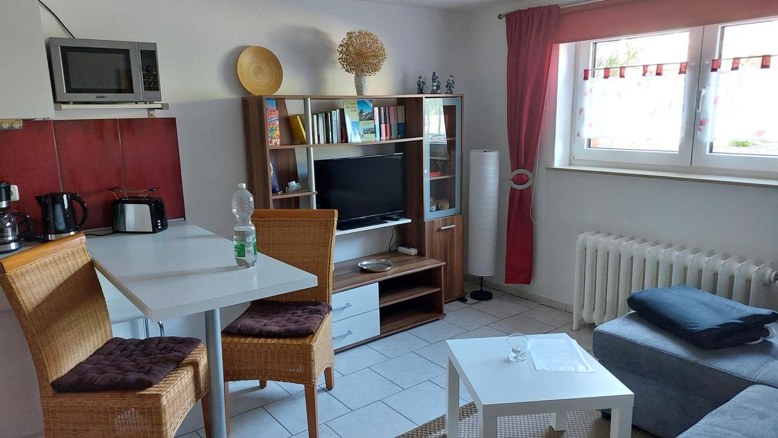 Wohn- und Essbereich mit Sofa, Tisch, Stühlen und Fernseher. Fenster mit roten Vorhängen.