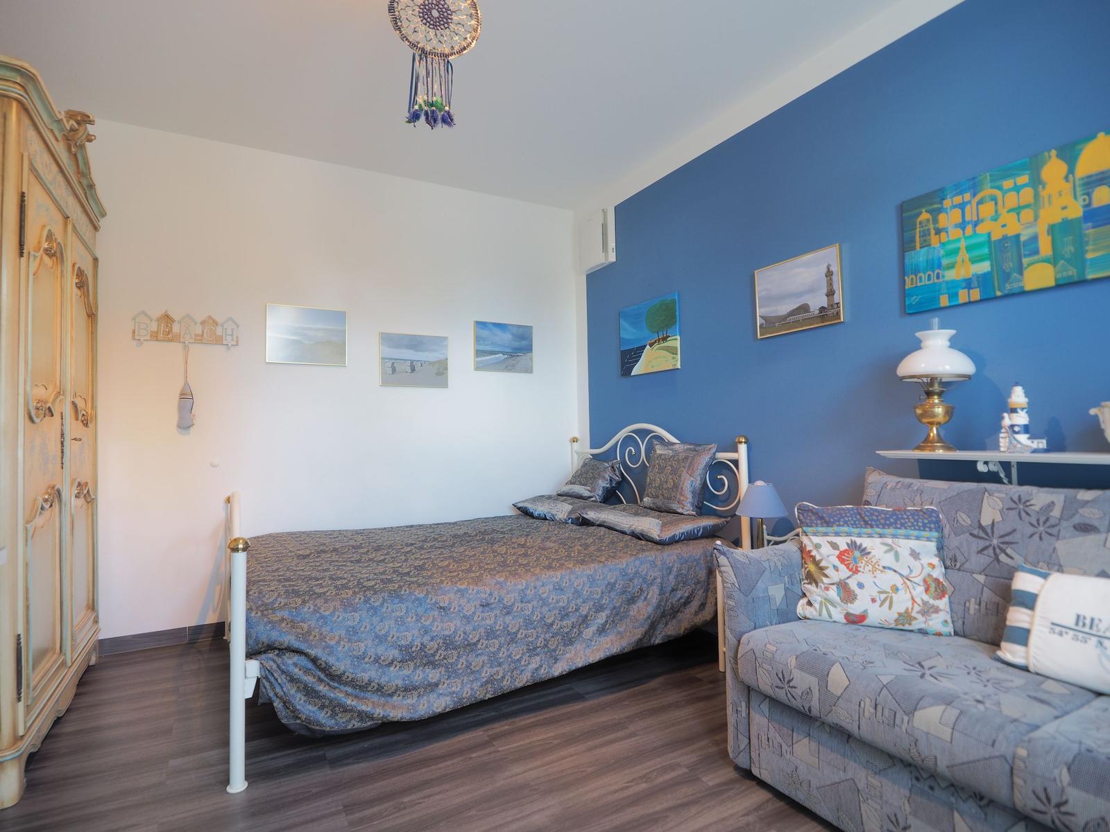 Schlafzimmer mit Bett, Sofa und Wandbildern. Ein Schrank und Decke sind sichtbar.